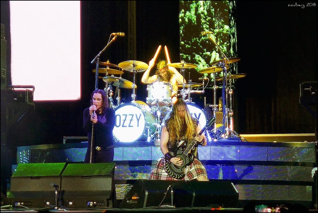 Ozzy Osbourne sings at a microphone stand in front of a bass drum reading OZZY, with a guitarist playing beside him and a drummer behind.