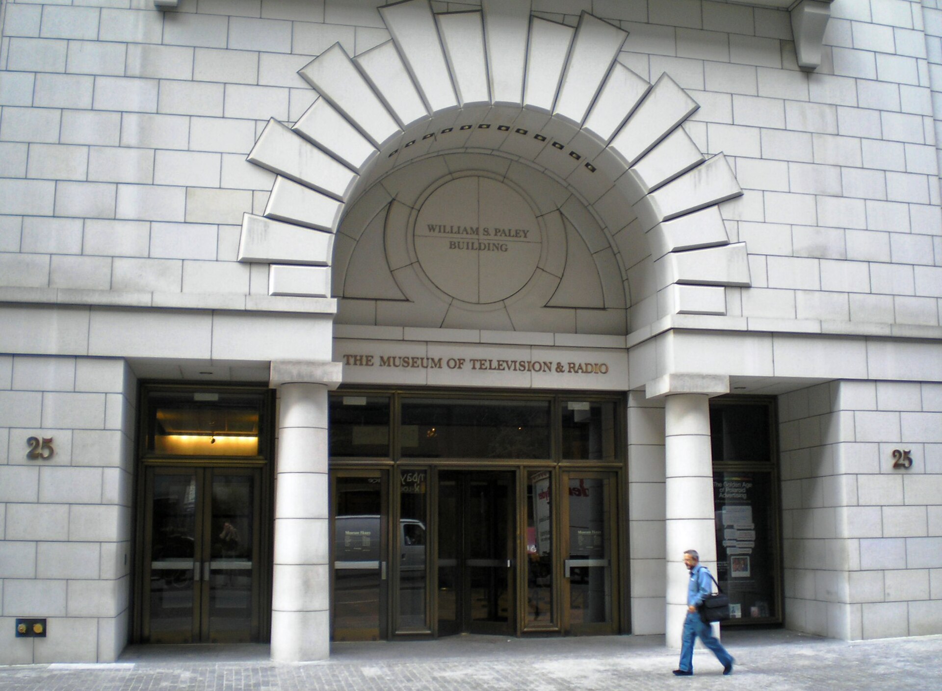 The limestone entrance of the William S. Paley Building in Manhattan, a fanned stone arch over brass doors, a man walking past.