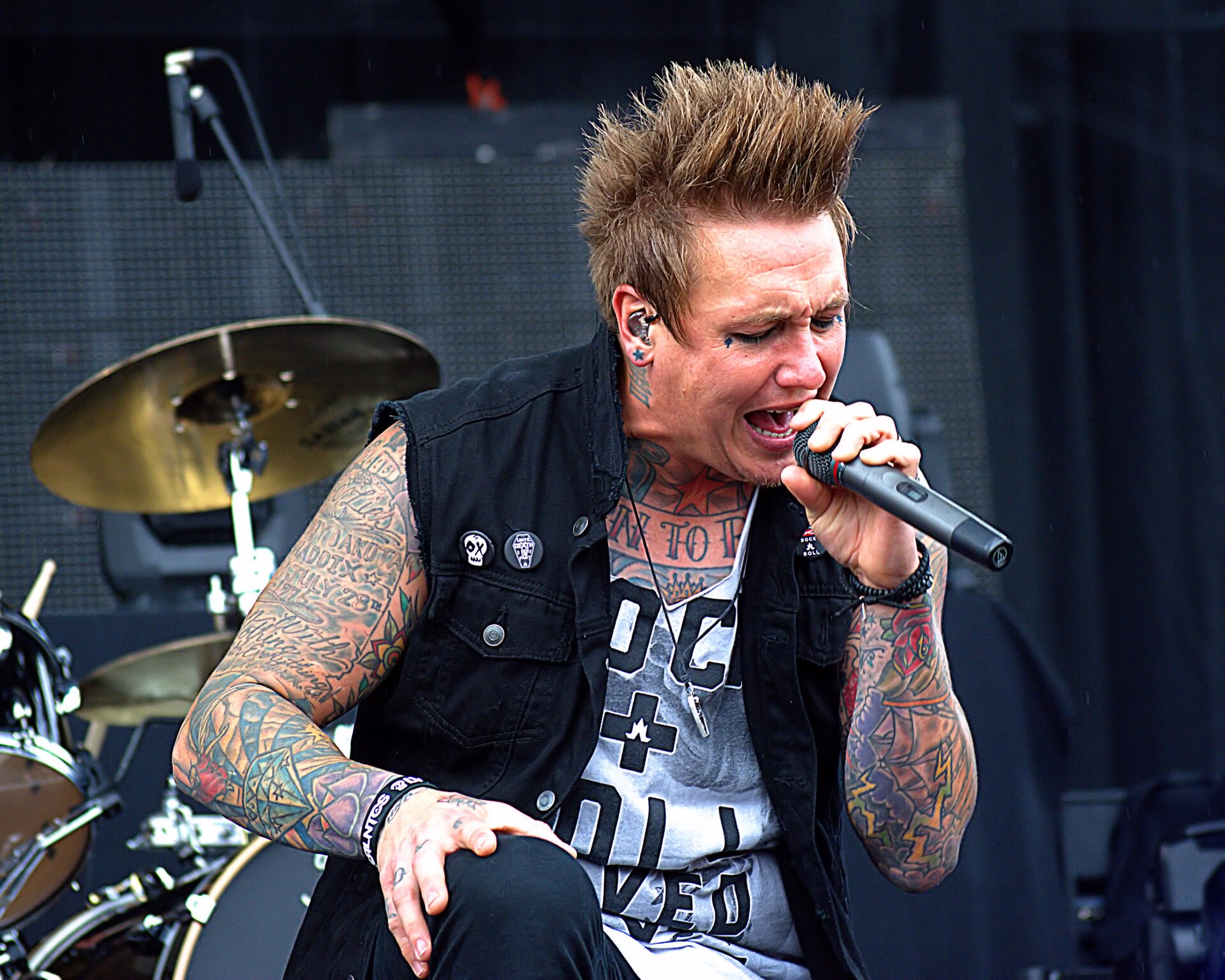 A heavily tattooed singer in a black cut-off vest screams into a handheld microphone on a festival stage, a drum kit behind him