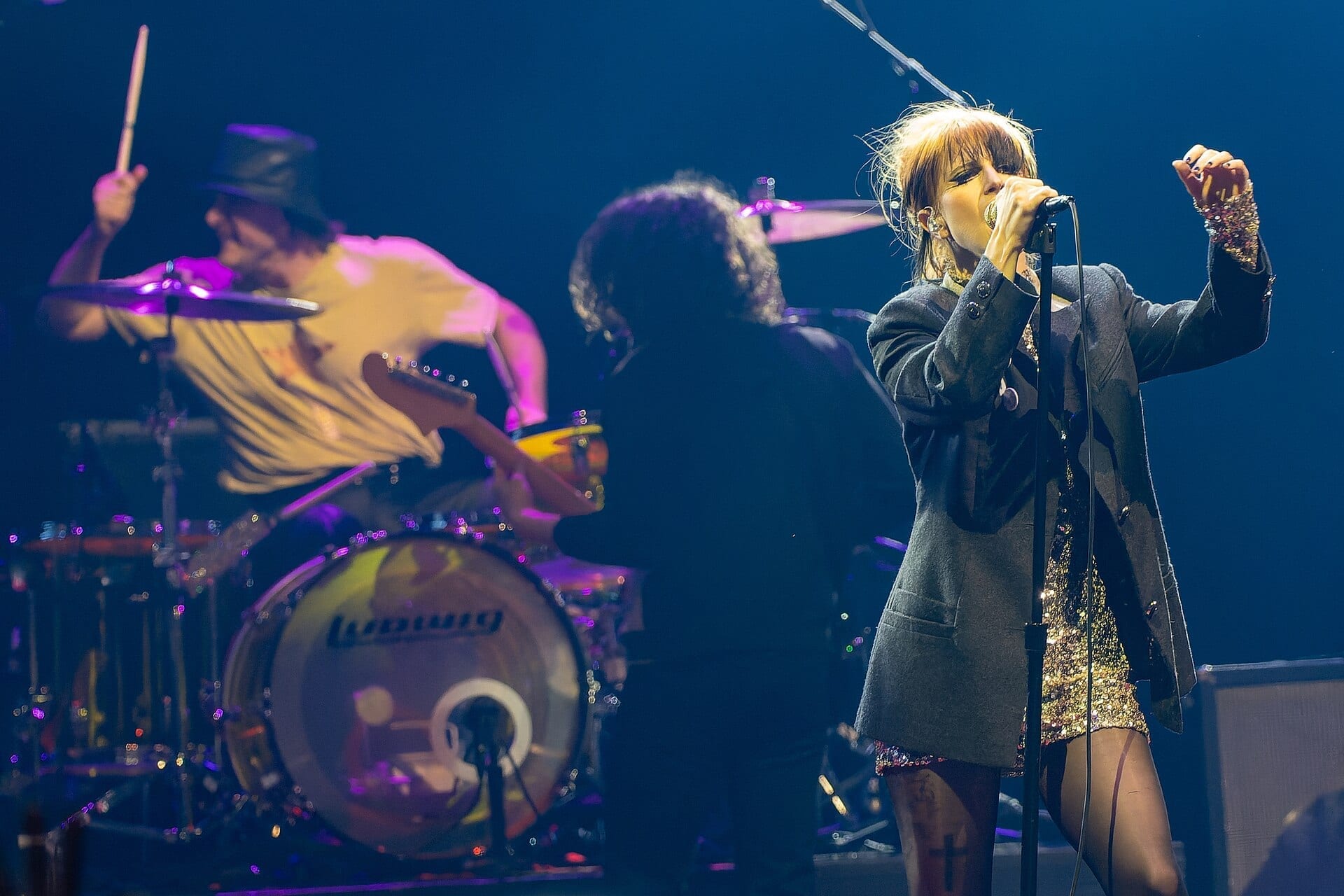 Paramore performing on stage: singer Hayley Williams in a sequined gold dress and gray coat sings into a microphone, with drummer Zac Farro in a top hat and guitarist Taylor York playing behind her.