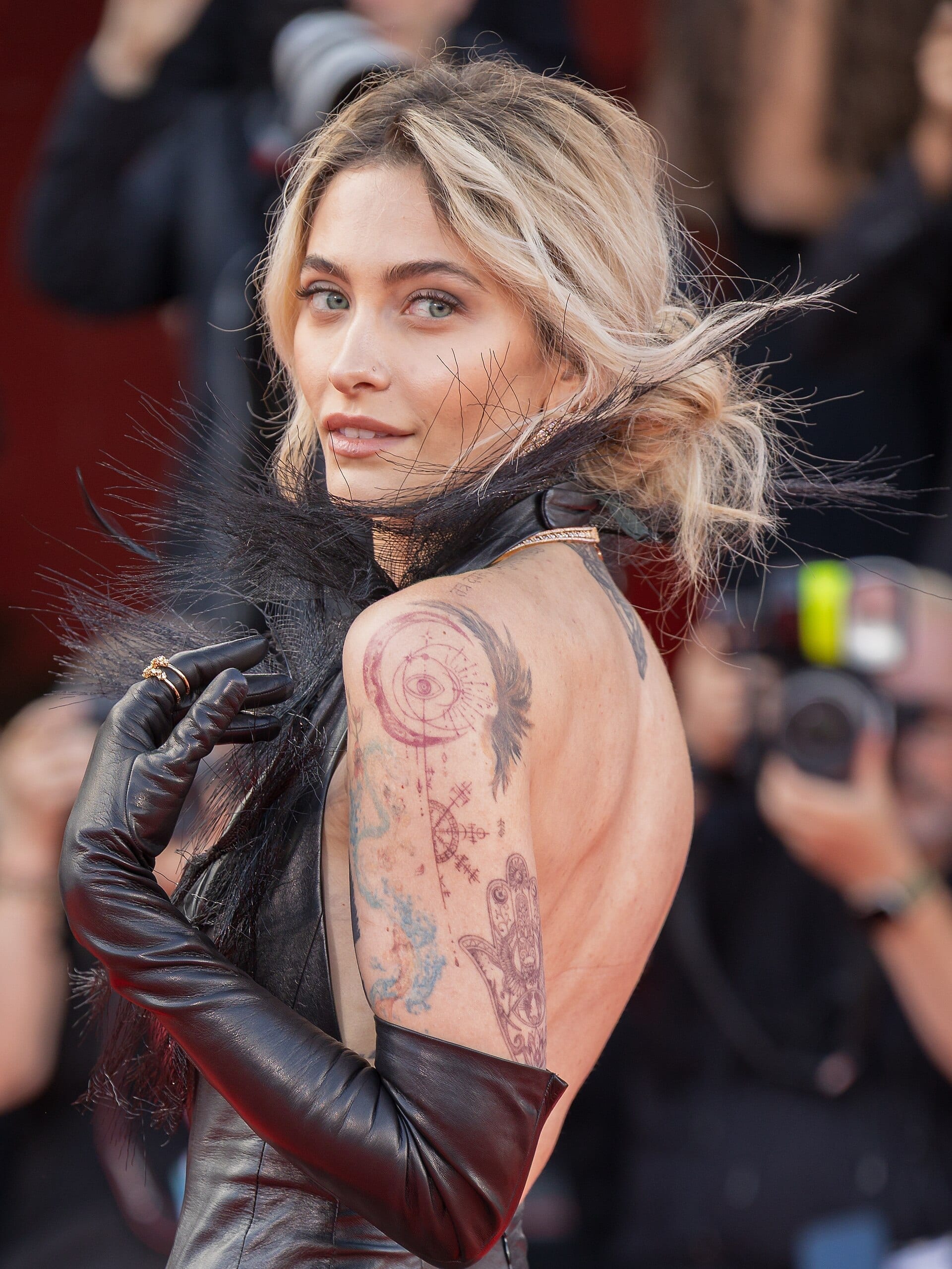 Paris Jackson looks back over a bare, tattooed shoulder in a black leather outfit with long gloves, her blonde hair loosely pinned up, on a red carpet.