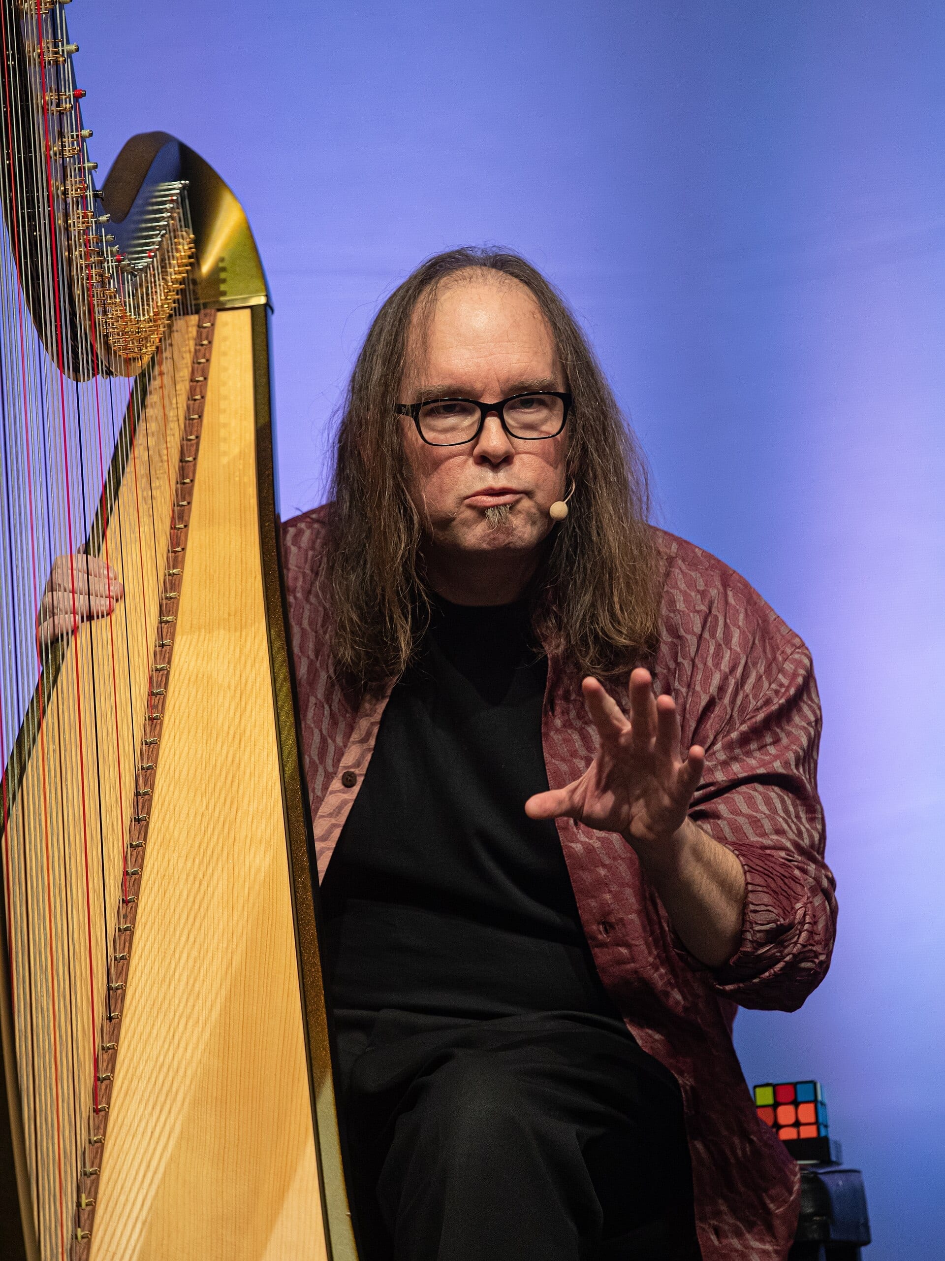 Park Stickney, wearing glasses and a patterned shirt, sits beside a large pedal harp with one hand resting on the strings and the other raised mid-gesture.