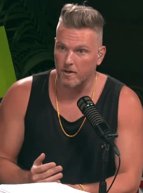 Pat McAfee, wearing a black tank top and a gold chain, speaks into a podcast microphone.