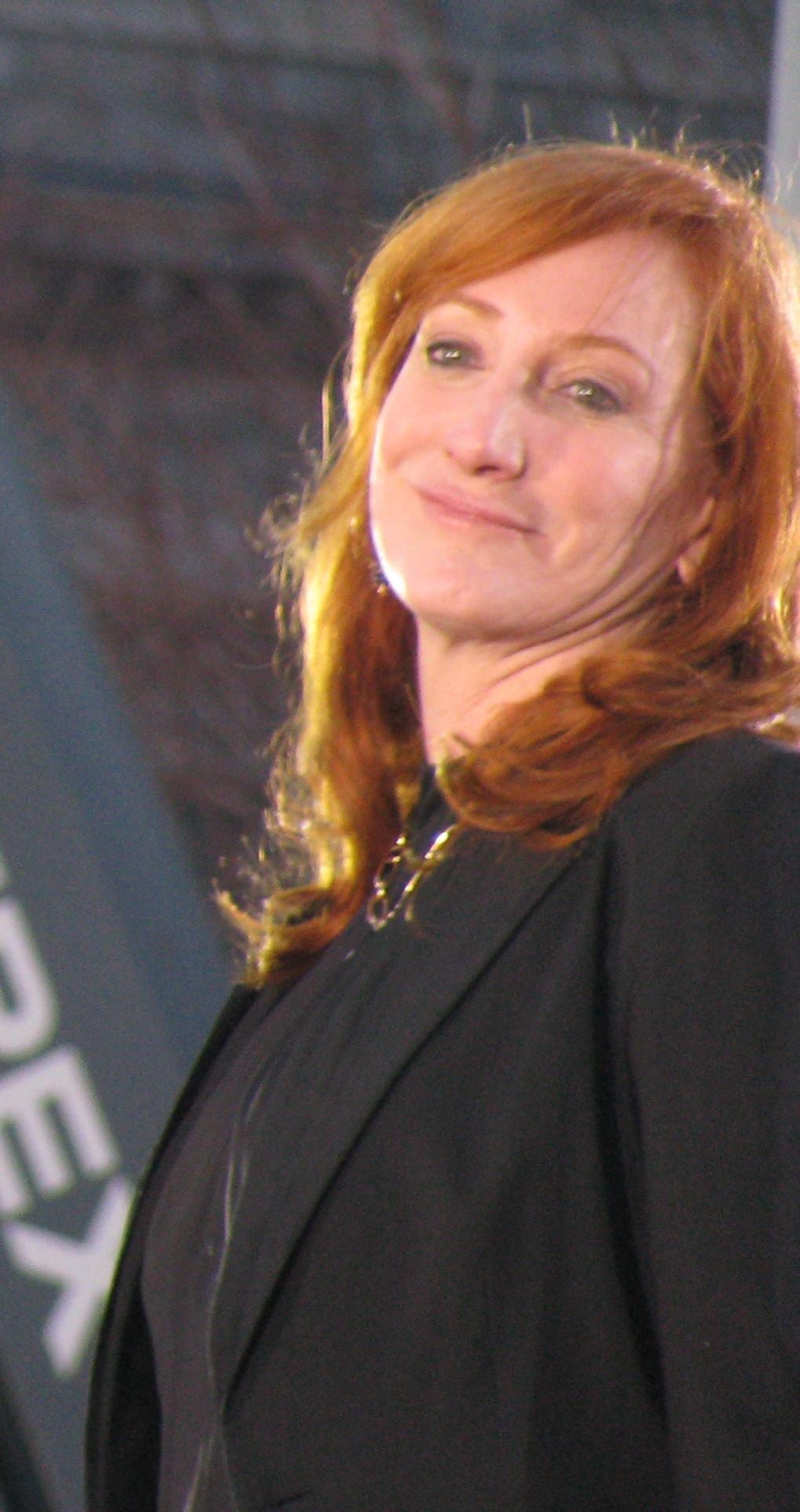 Patti Scialfa smiles while tilting her head, wearing a black jacket, with her red hair falling loose.