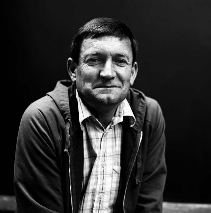 Black and white portrait of Paul Heaton, half-smiling straight at the camera in a zipped hooded top over a checked shirt, against a dark background.