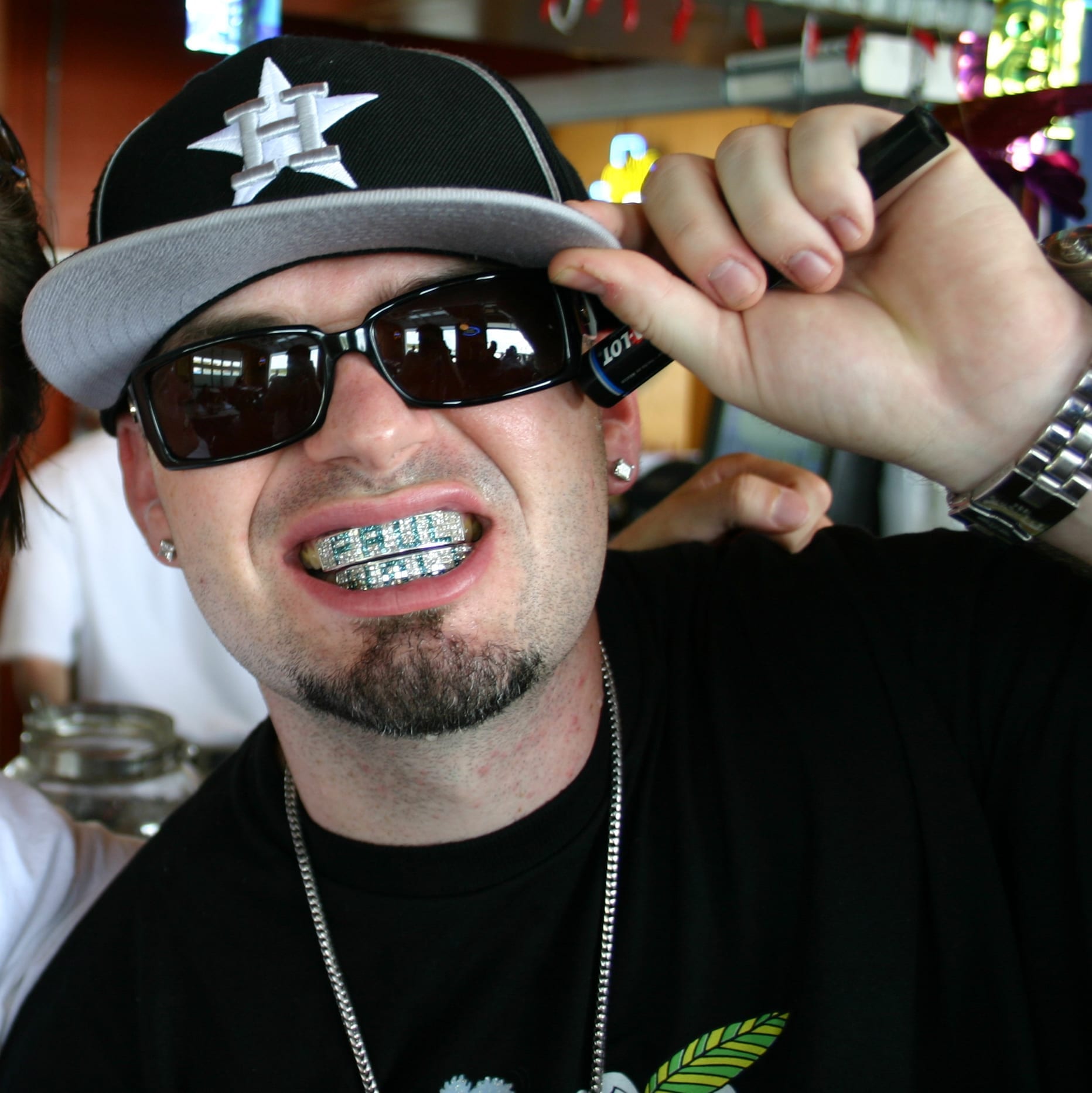 Paul Wall, wearing sunglasses and a baseball cap, smiles to show off his diamond-encrusted grill while tipping his cap with one hand.