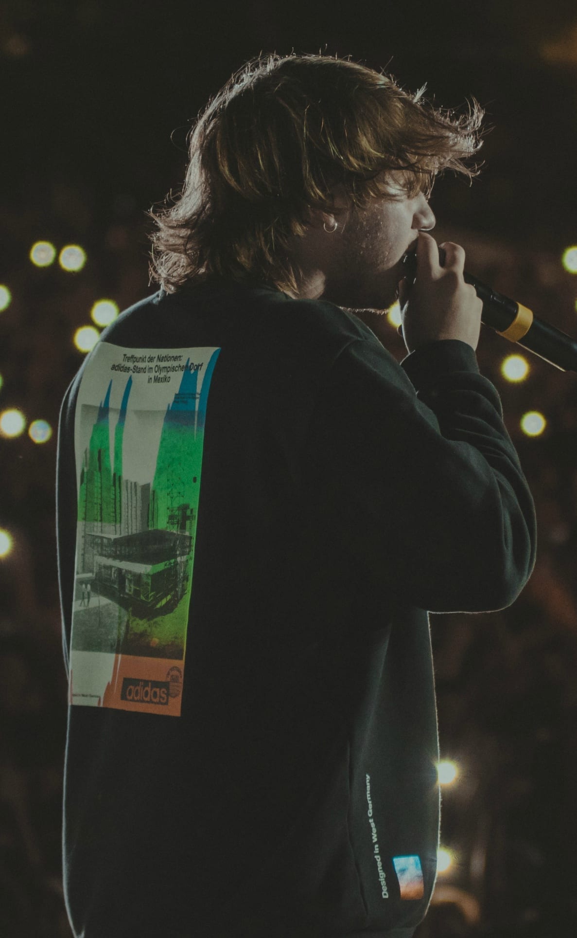 Paulo Londra sings into a microphone in profile, his hair blown back, wearing a dark jacket printed with a vintage Adidas graphic, lit against a dark concert backdrop.