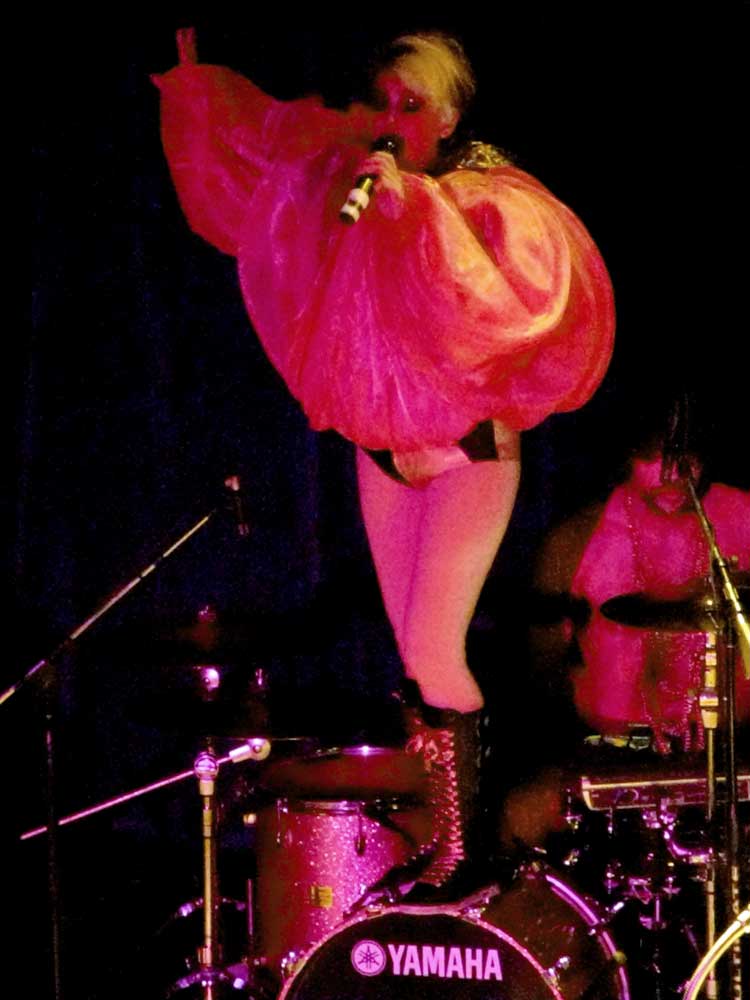 Peaches stands on a bass drum in an enormous pink ruffled costume, singing into a handheld microphone under pink light.