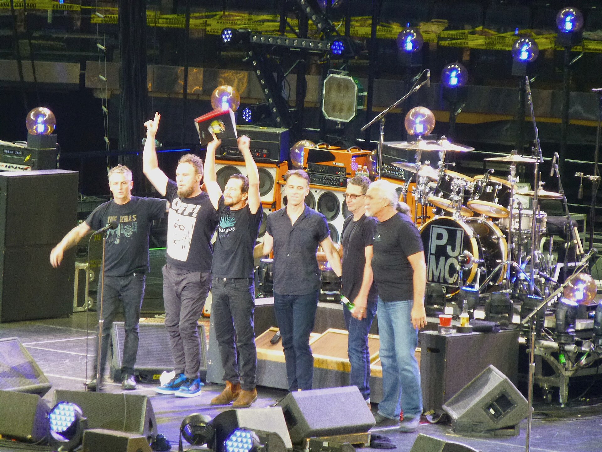 The members of Pearl Jam stand in a line at the front of an arena stage, one holding a record sleeve overhead, a bass drum reading PJ MC behind them.