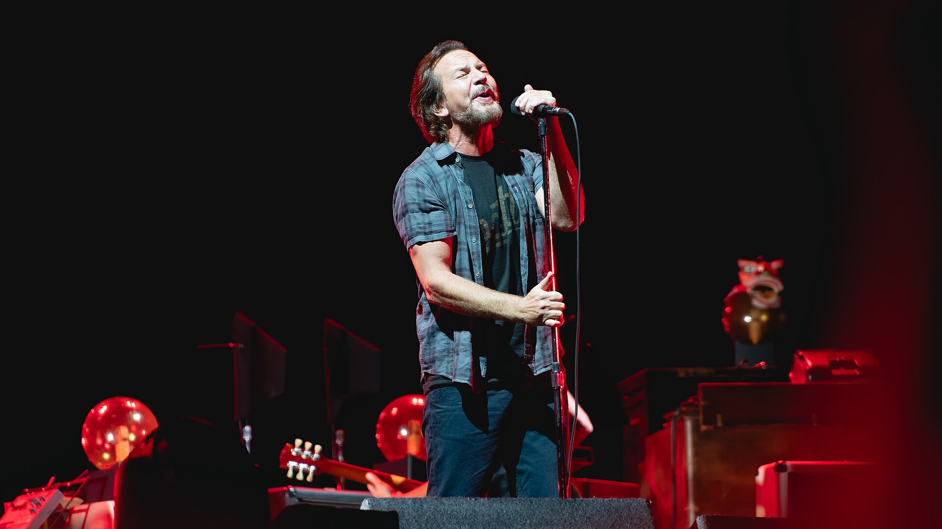 Eddie Vedder sings with both hands on a microphone stand under red stage light, a plaid shirt open over a black T-shirt.