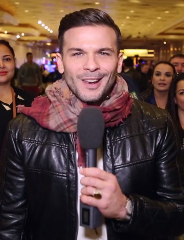 Pedro Capó speaking into an interview microphone in a black leather jacket and a red plaid scarf, a crowded hotel lobby behind him