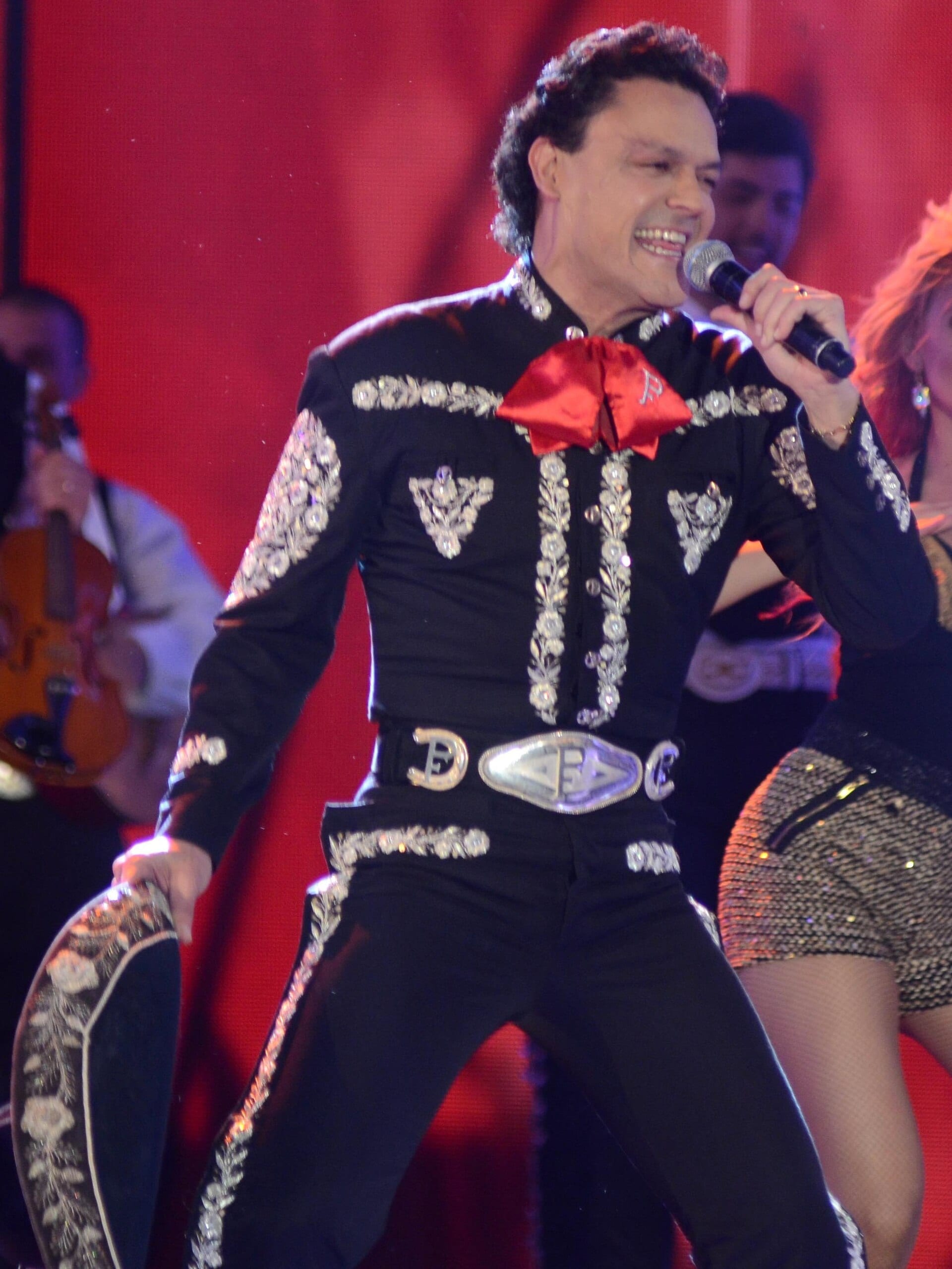 Pedro Fernandez sings into a microphone on stage, wearing a black charro suit with silver embroidery and a red bow tie monogrammed with the letter P.