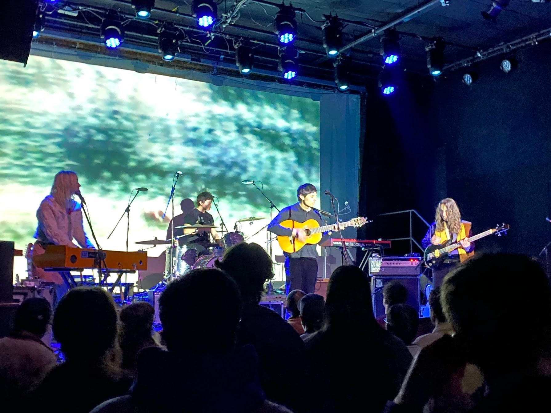 Peel Dream Magazine play a club stage seen over the heads of the audience, four musicians on keys, drums, nylon-string guitar and bass in front of a projected film of leaves.