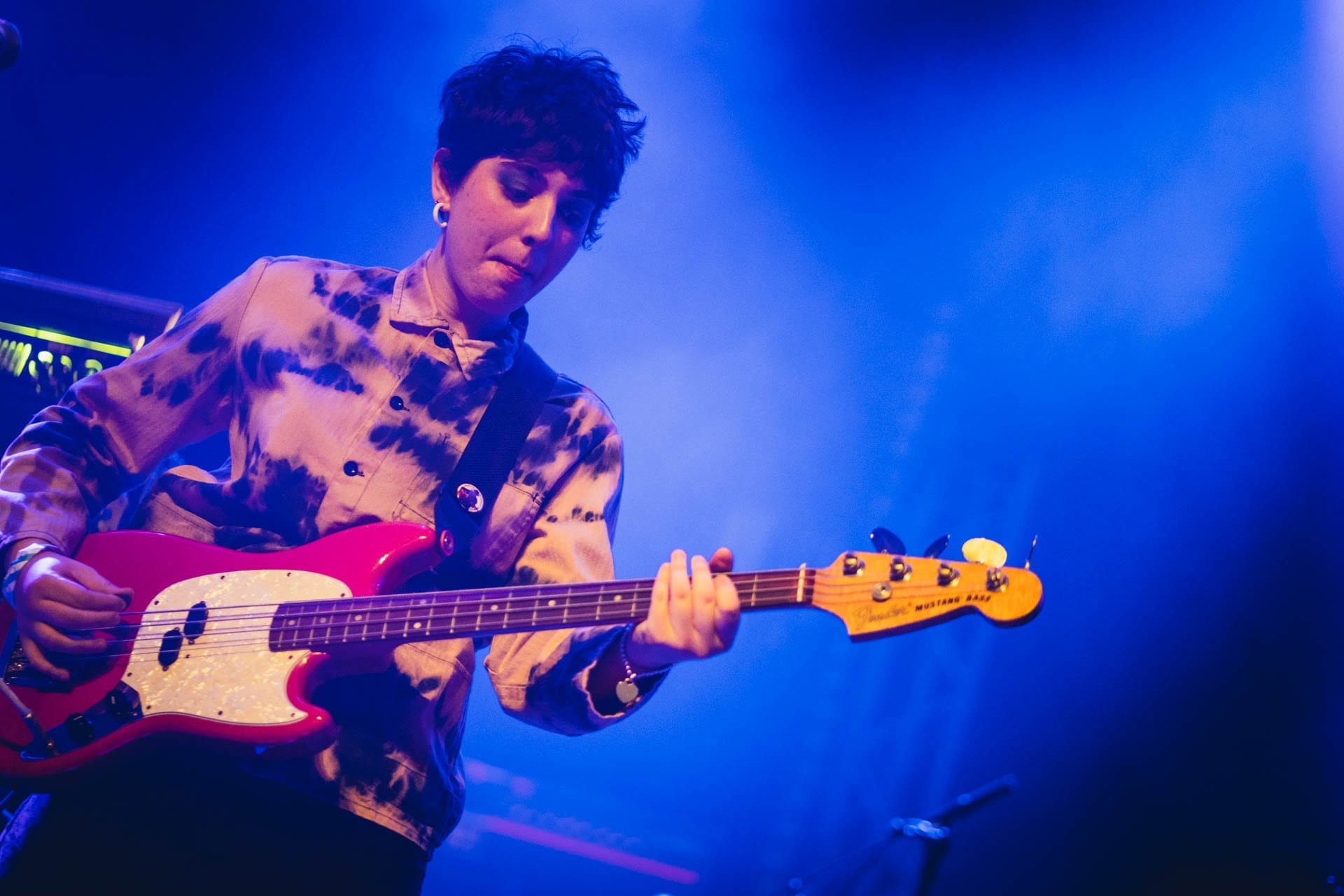 A member of Penelope Isles, wearing a tie-dyed jacket, plays a pink Fender bass guitar under deep blue stage light.