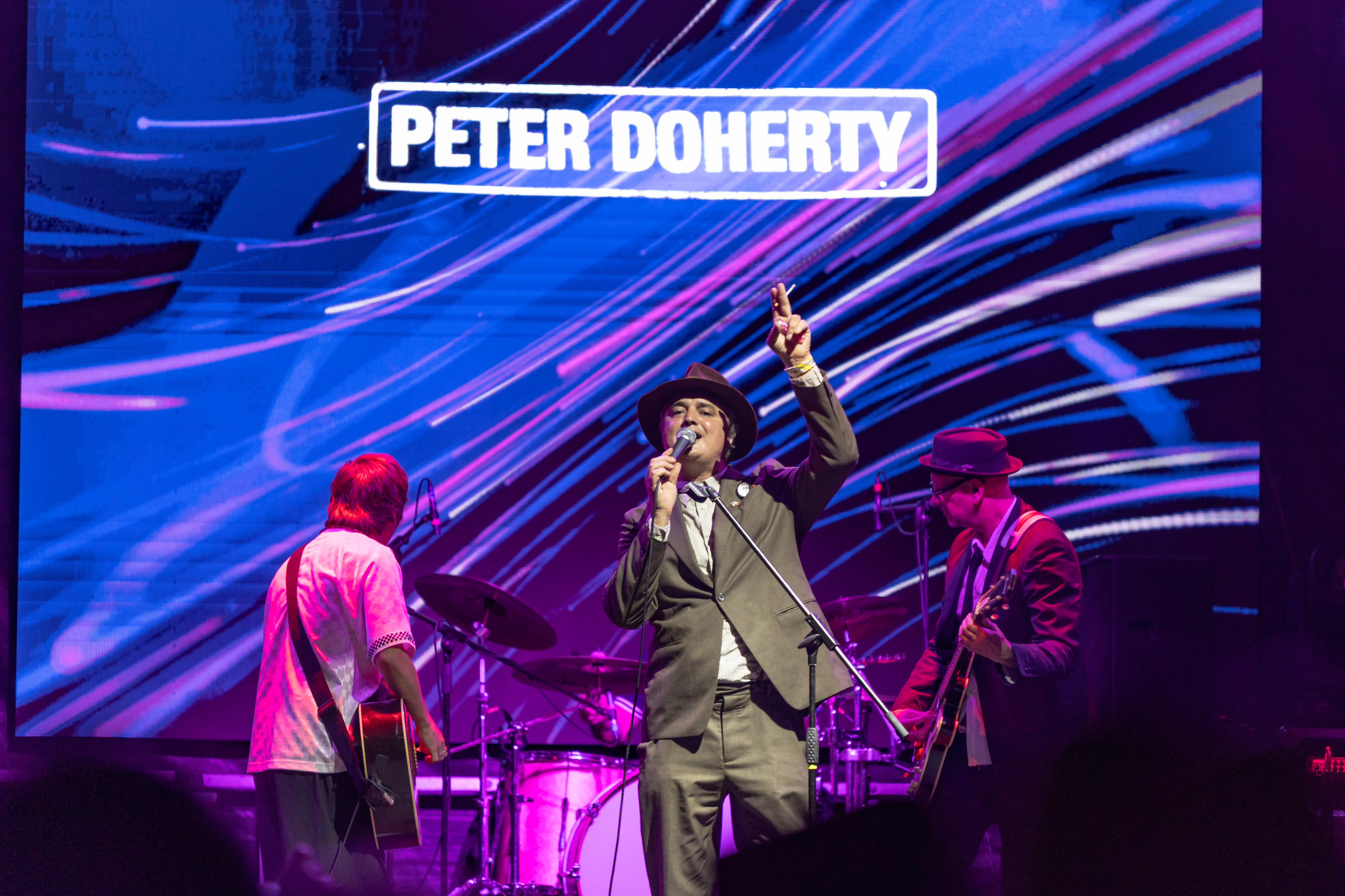 Pete Doherty sings at a microphone stand in a hat and suit with one hand raised, his name lit on the screen behind him and a guitarist either side.