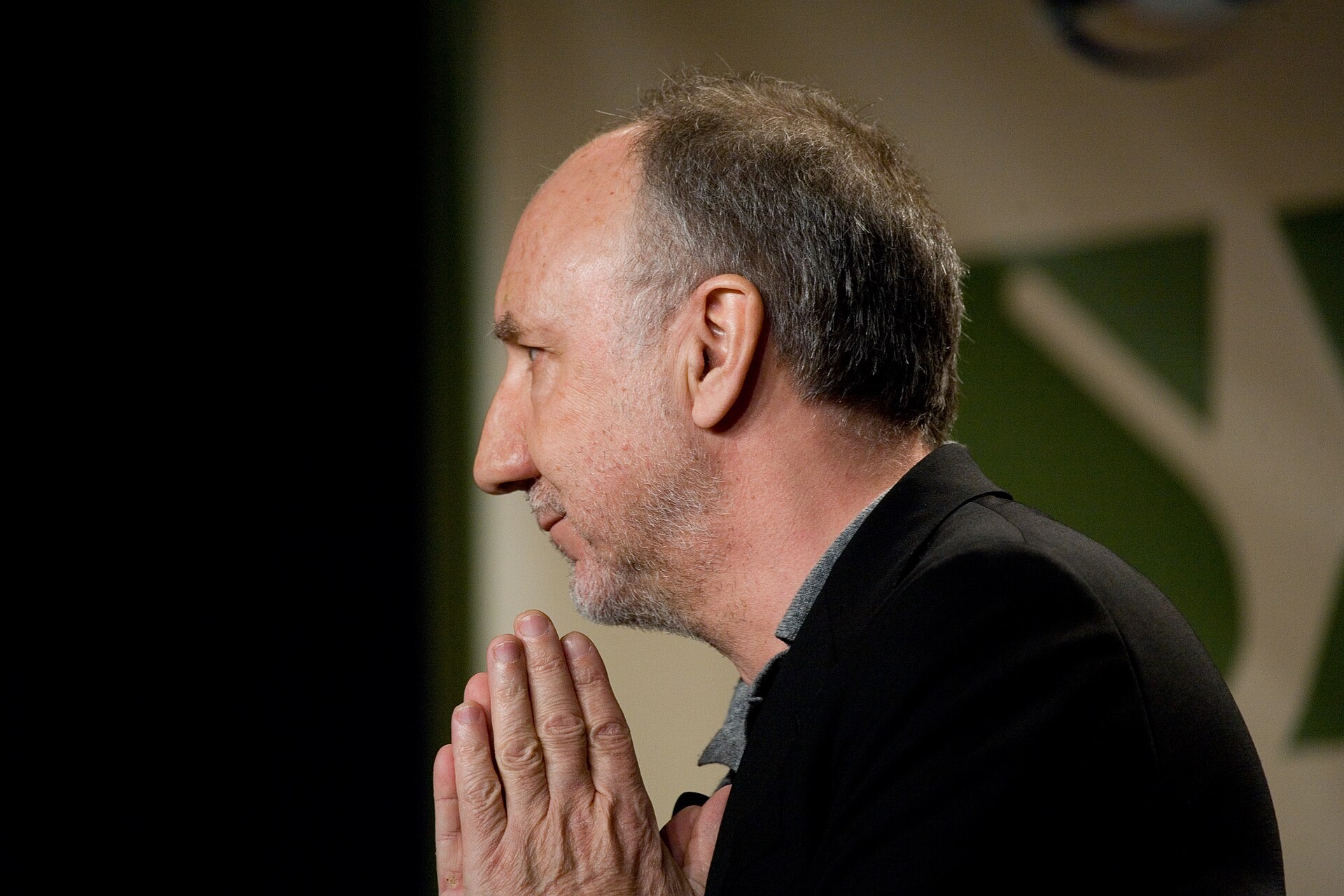 Pete Townshend in profile against a dark background, greying hair swept back and hands pressed together in front of his chin, wearing a black jacket over a grey collar.