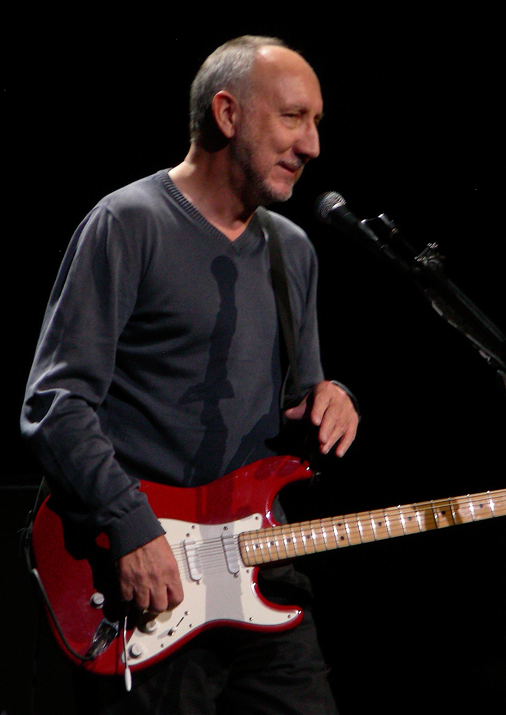 Pete Townshend plays a red Fender Stratocaster at a microphone, grey jumper, half smiling.