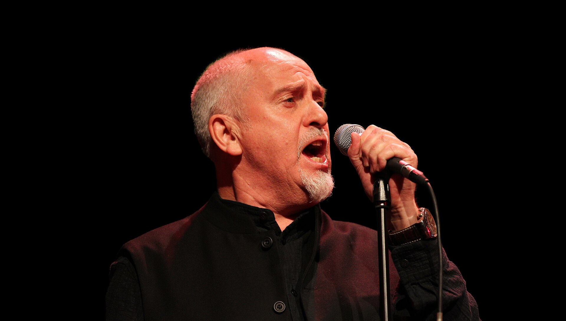 Peter Gabriel sings with his eyes closed, close-shaven head and a white goatee, in a dark jacket against a black background.