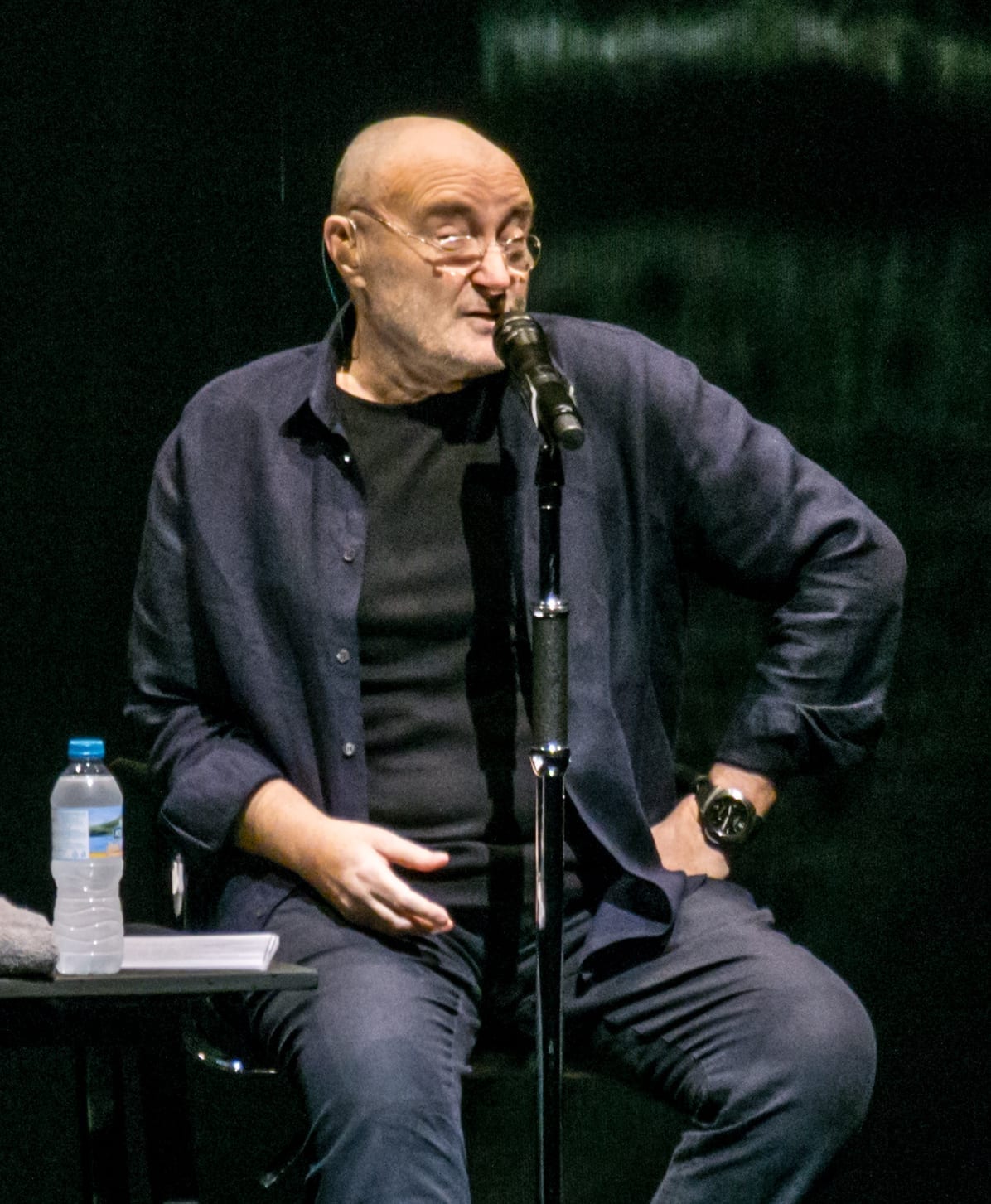 Phil Collins, seated on stage in a dark shirt and jacket, speaks into a microphone stand with a water bottle on a side table.