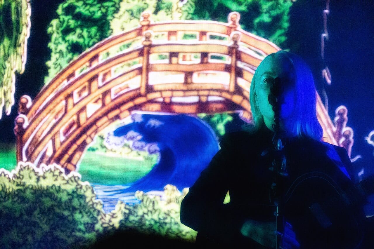 Phoebe Bridgers sings into a microphone under blue stage light, in front of a large screen showing an illustrated arched wooden bridge over water.