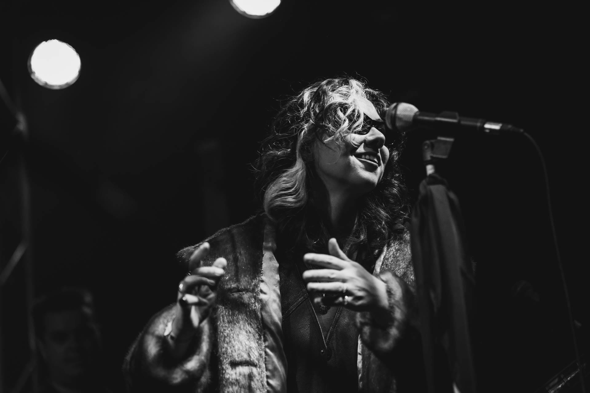 Black and white photo of a singer in a fur-collared coat, hands raised, leaning back from a microphone on a small stage.