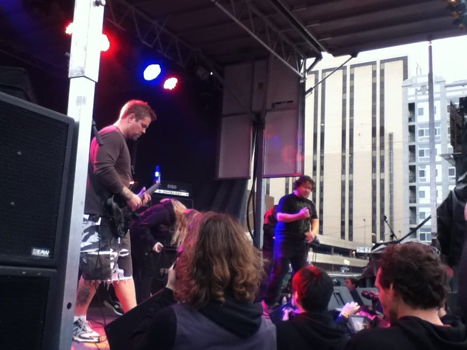 Two members of Pig Destroyer perform on an outdoor daytime festival stage, one playing guitar, with a crowd watching and city buildings visible behind the stage.