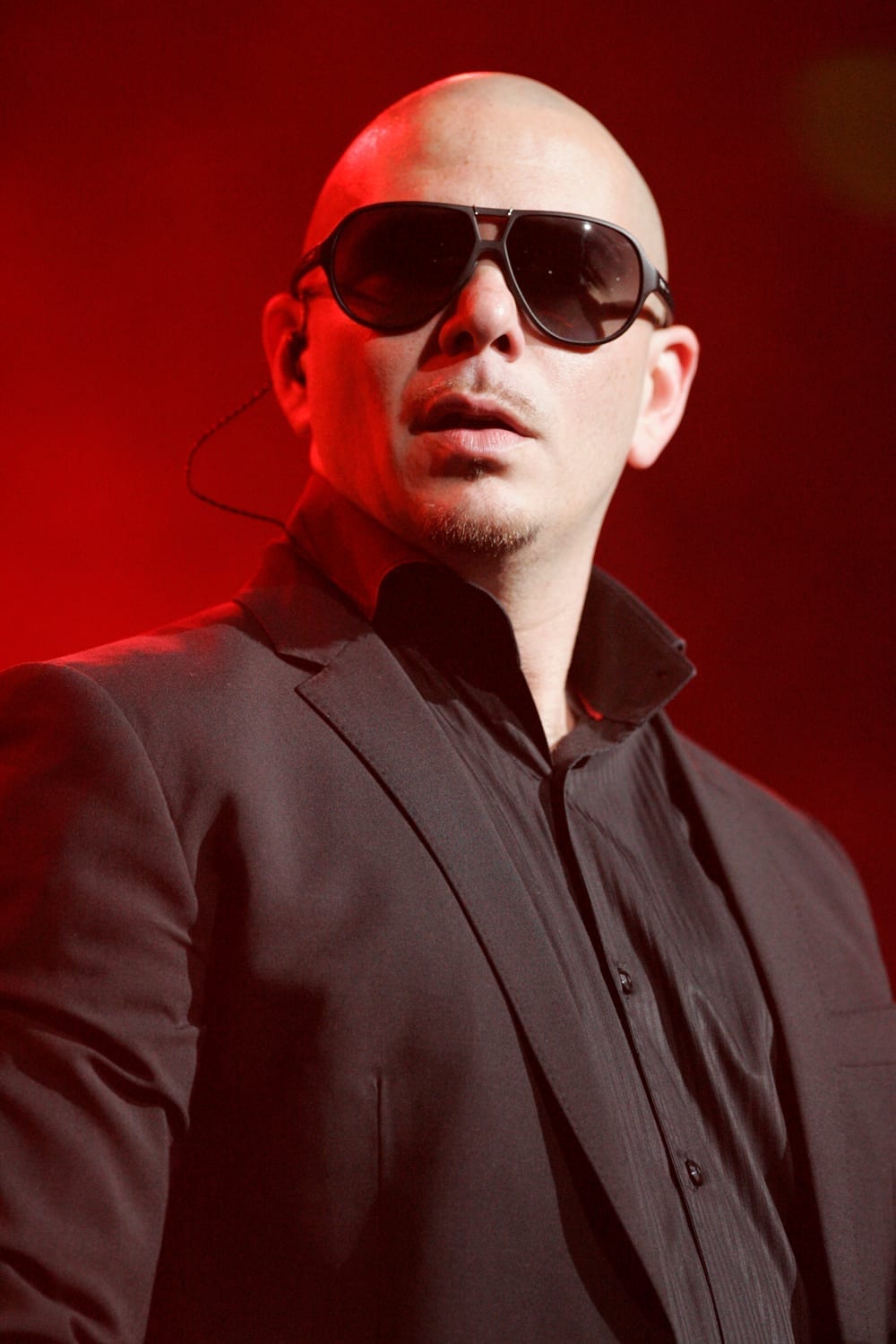 Pitbull standing on stage in a black suit and aviator sunglasses, lit by red stage lighting.