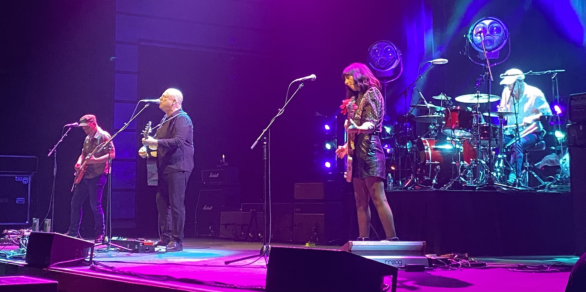 The Pixies play a wide stage under magenta light, with two guitarists at microphones on the left and the drum kit raised behind them.