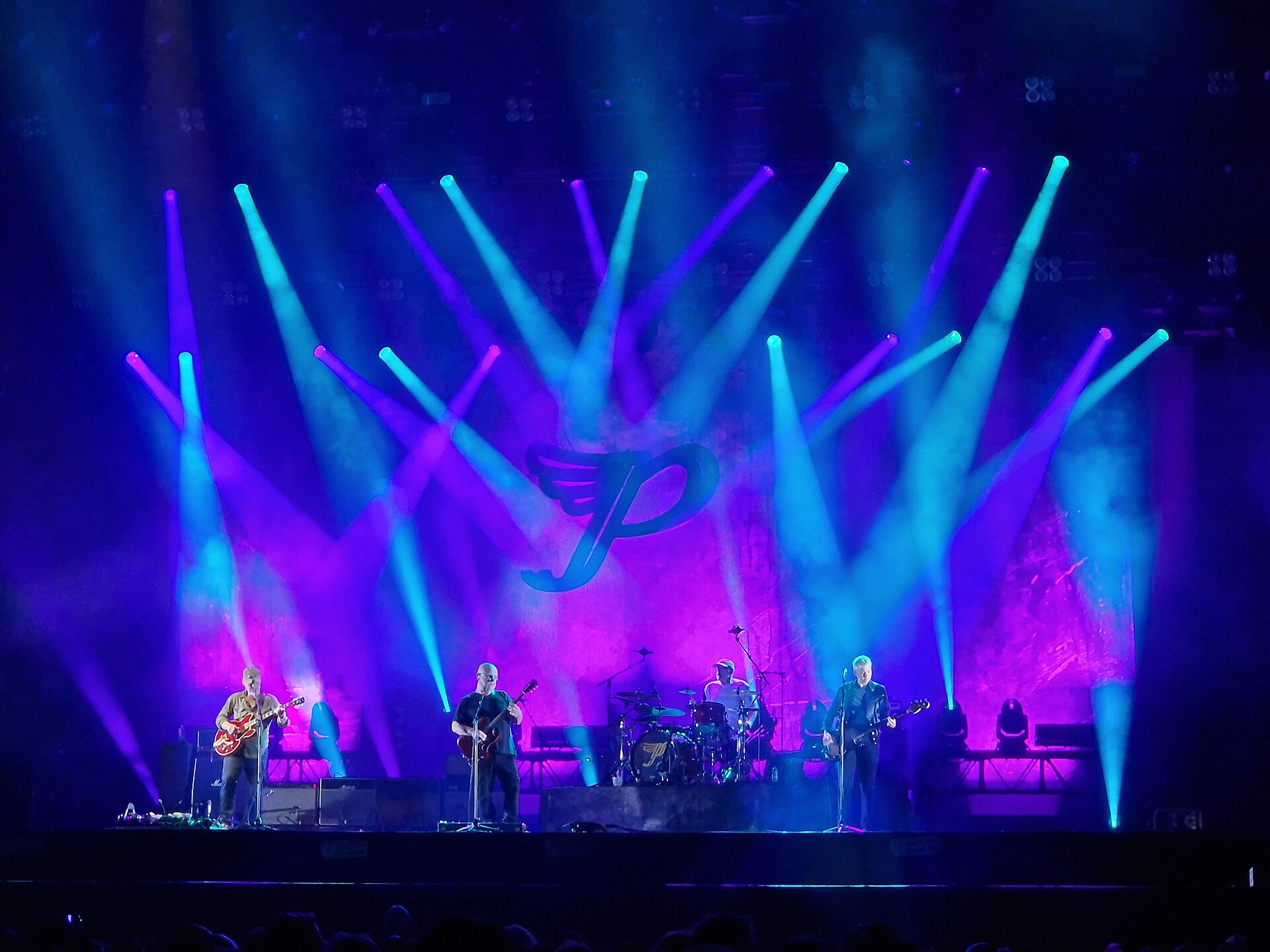 Pixies play a festival stage under a fan of blue and purple beams, the band's winged P logo lit on the backdrop behind them.