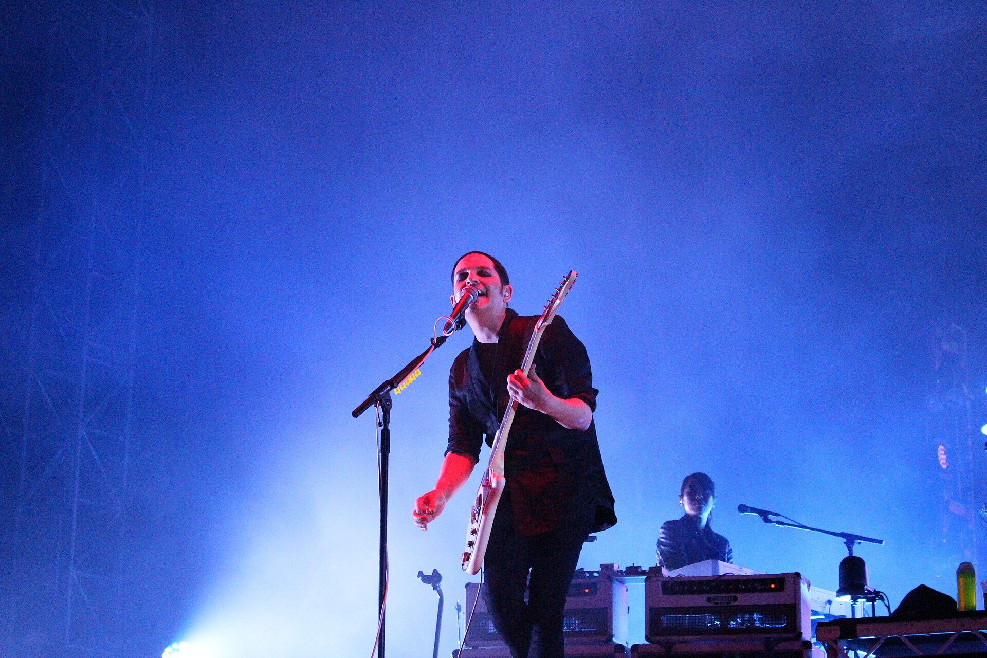 Brian Molko of Placebo singing into a mic stand while playing guitar, lit red against a wall of blue stage haze, a keyboard player behind him