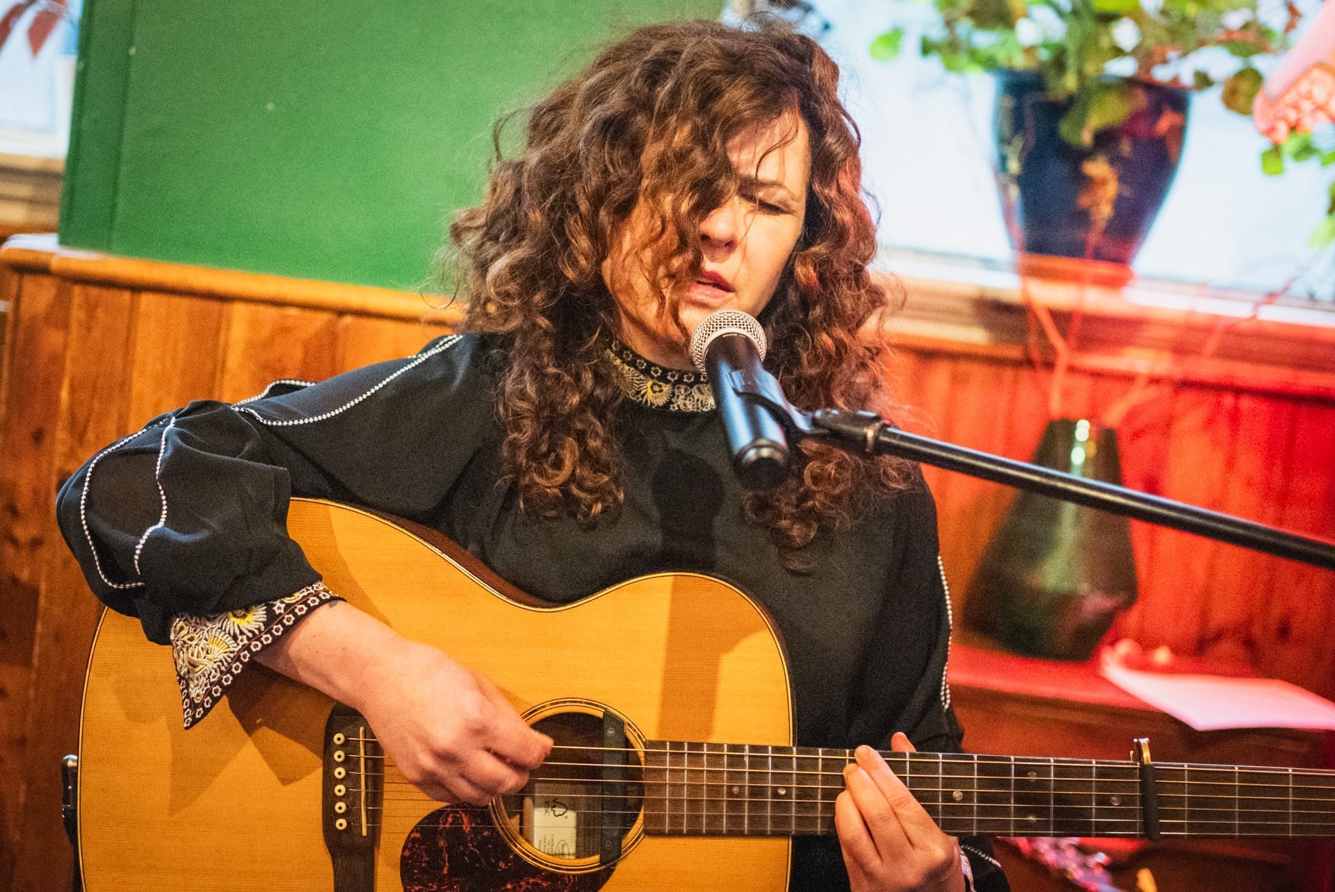 Polly Paulusma sings into a microphone while playing an acoustic guitar, eyes closed, wearing an embroidered dark blouse.
