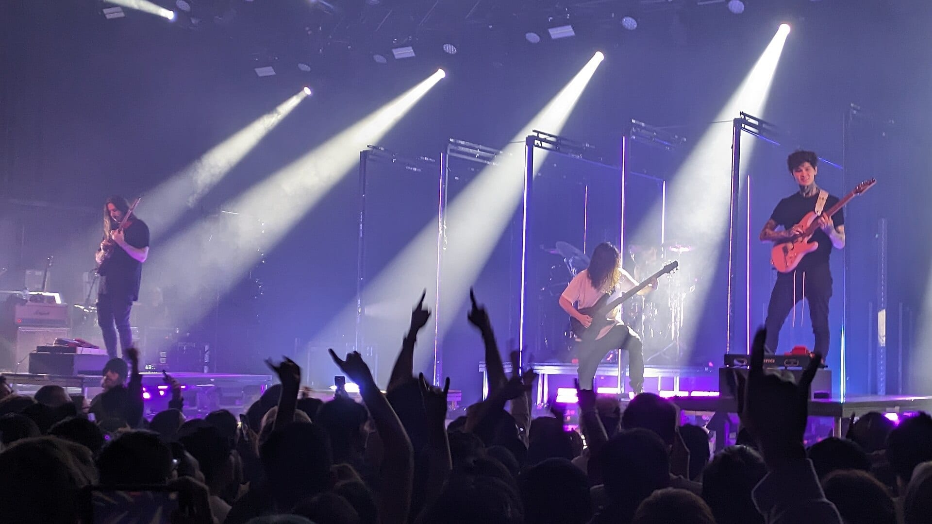 Polyphia spread across a smoky stage lit by four hard white beams, two guitarists on risers at either side and a third crouched at the center, hands raised in the crowd below.