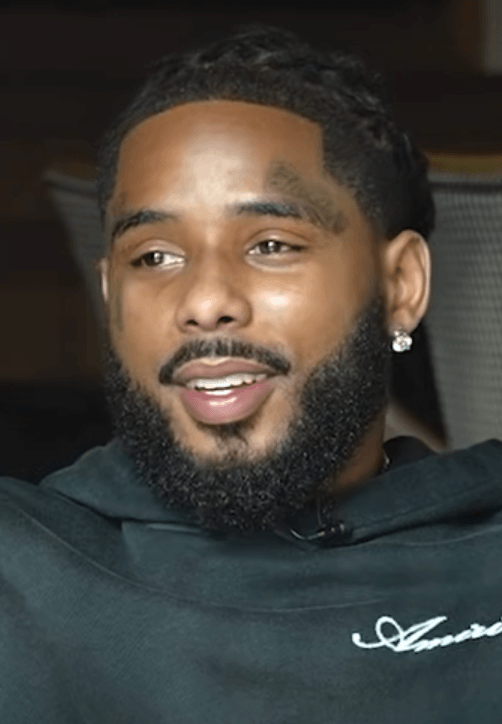 Pooh Shiesty smiles mid-sentence during an interview, wearing a dark hoodie, with a full beard and a small tattoo above one eyebrow.