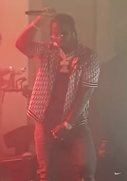 Pop Smoke performs on stage, raising a microphone above his head with one hand, wearing a patterned varsity-style jacket under red stage lighting.