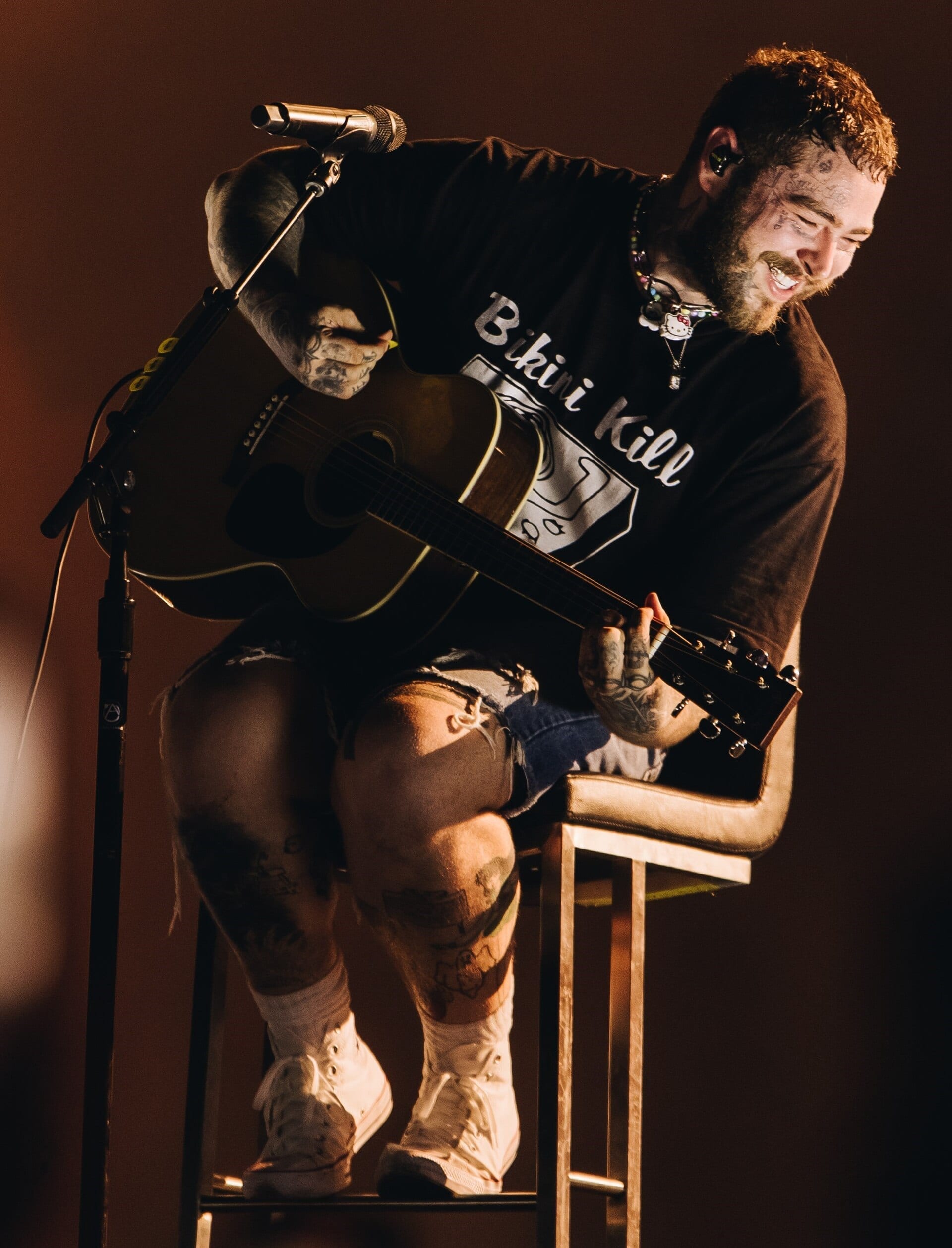 Post Malone laughs while playing an acoustic guitar on a bar stool, in a Bikini Kill T-shirt and cut-off denim shorts, tattoos across his face and legs.