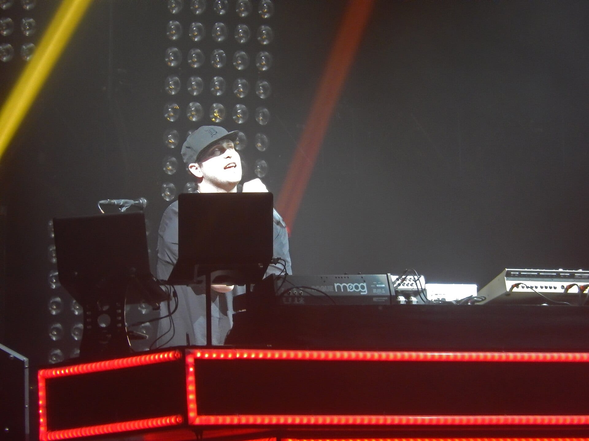 Pretty Lights looks up from behind a Moog and a laptop stand in a baseball cap, the booth edged in red bulbs and a wall of lights behind him.