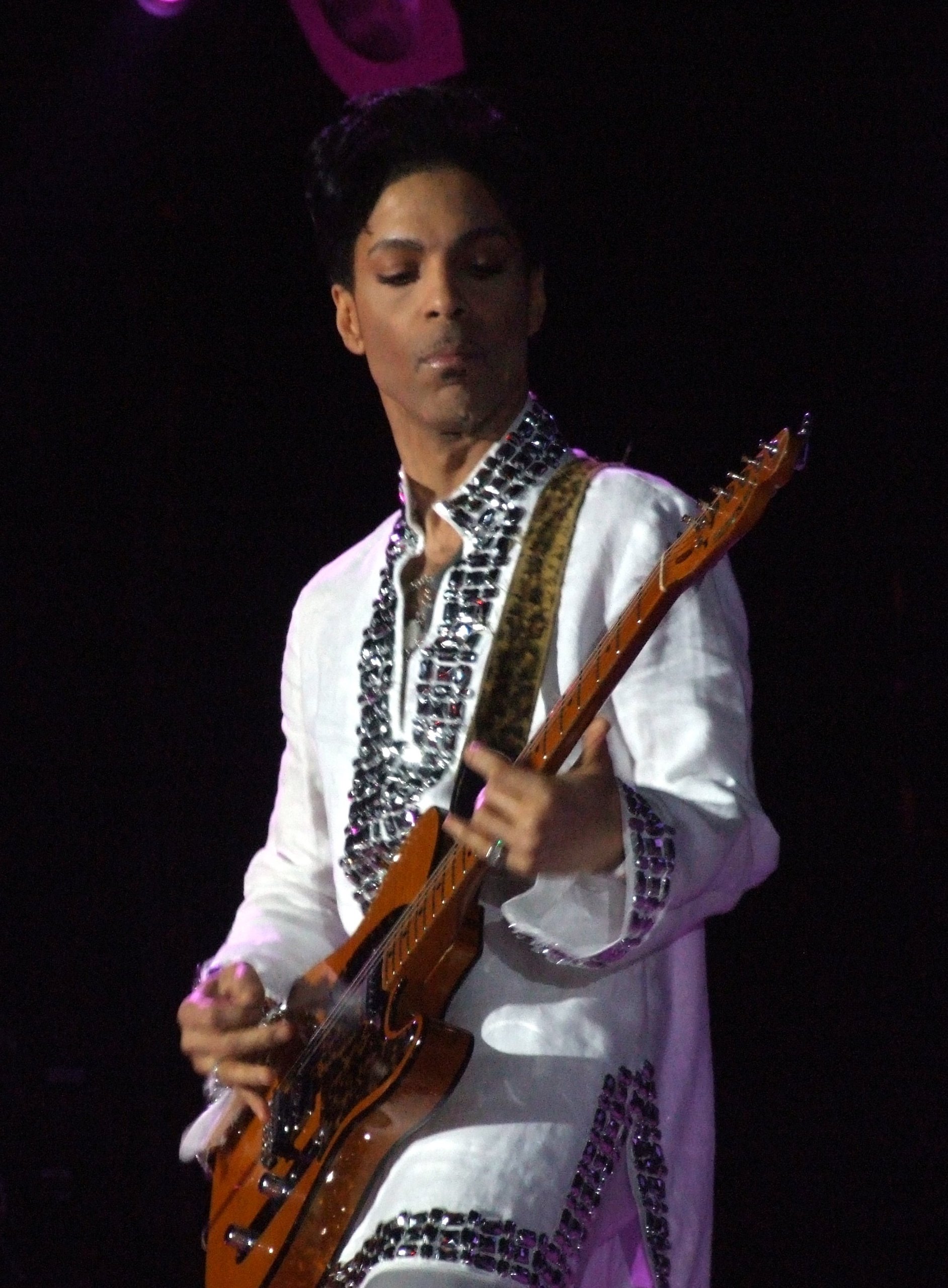 Prince plays a white electric guitar on a night stage in a white jacket and a wide-brimmed hat, lit from behind.