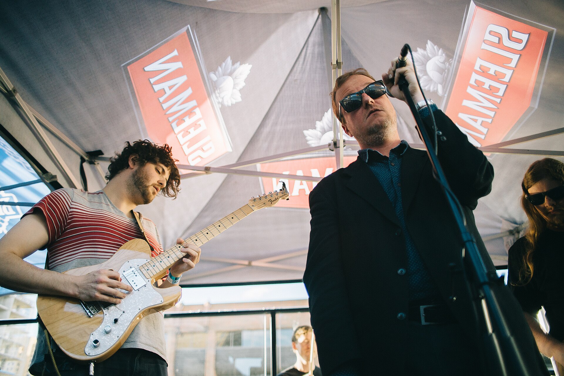 Protomartyr's singer tips a microphone stand toward himself in sunglasses and a dark blazer while a guitarist plays a Telecaster beside him under a canopy.