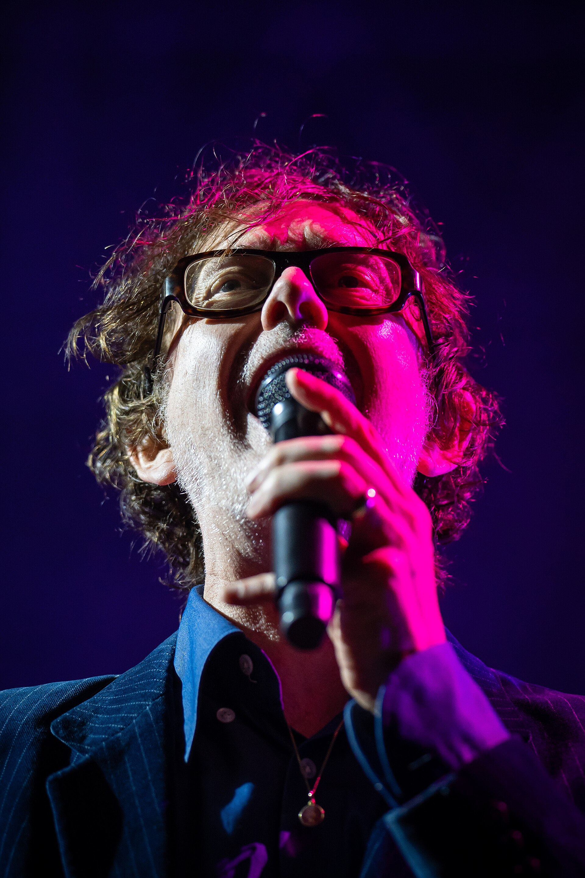 Jarvis Cocker sings into a handheld microphone in pink light, wearing heavy dark glasses and a jacket over a shirt.