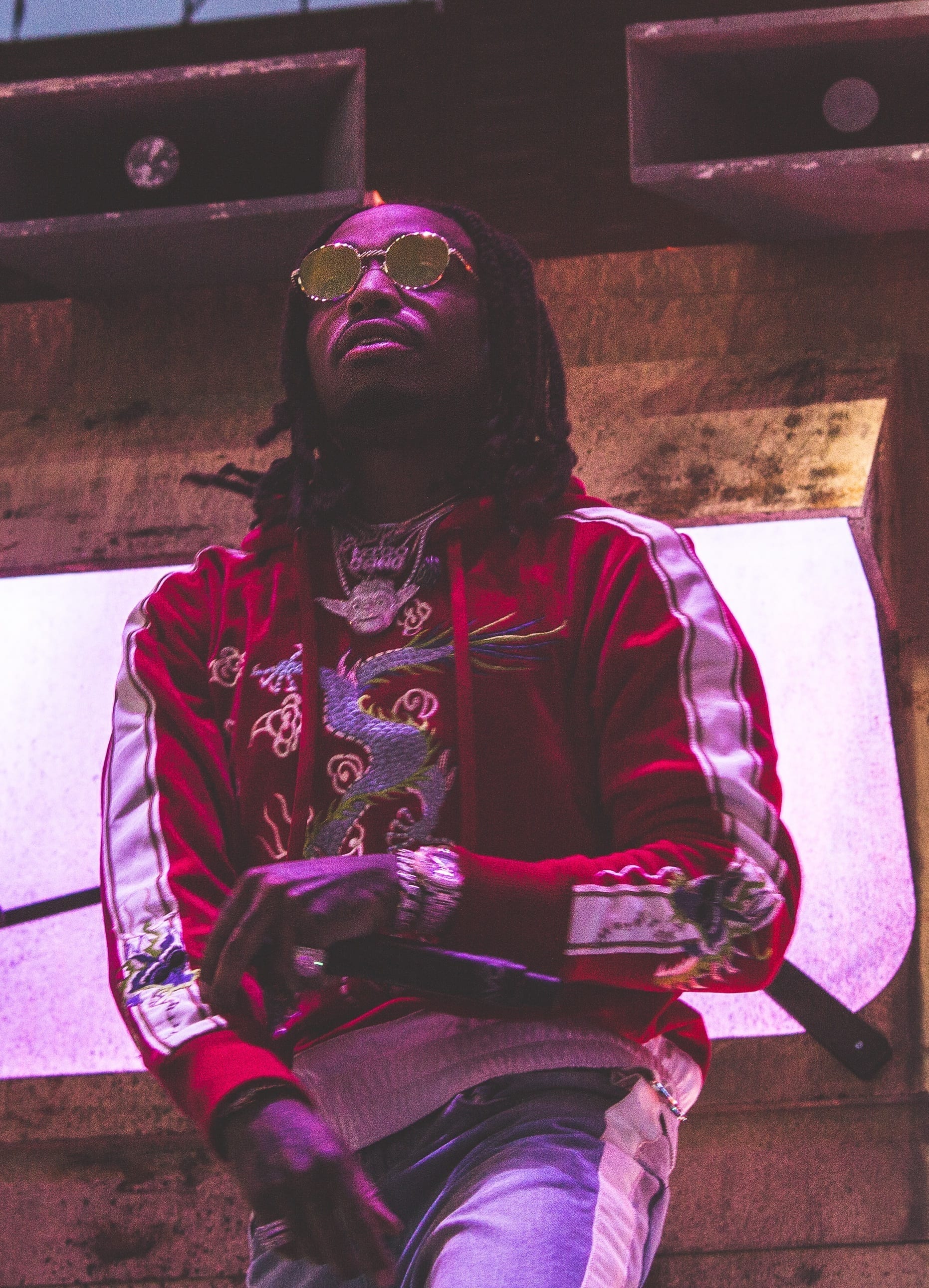 Quavo, wearing round tinted sunglasses and a red dragon-embroidered jacket, looks upward under purple stage lighting.