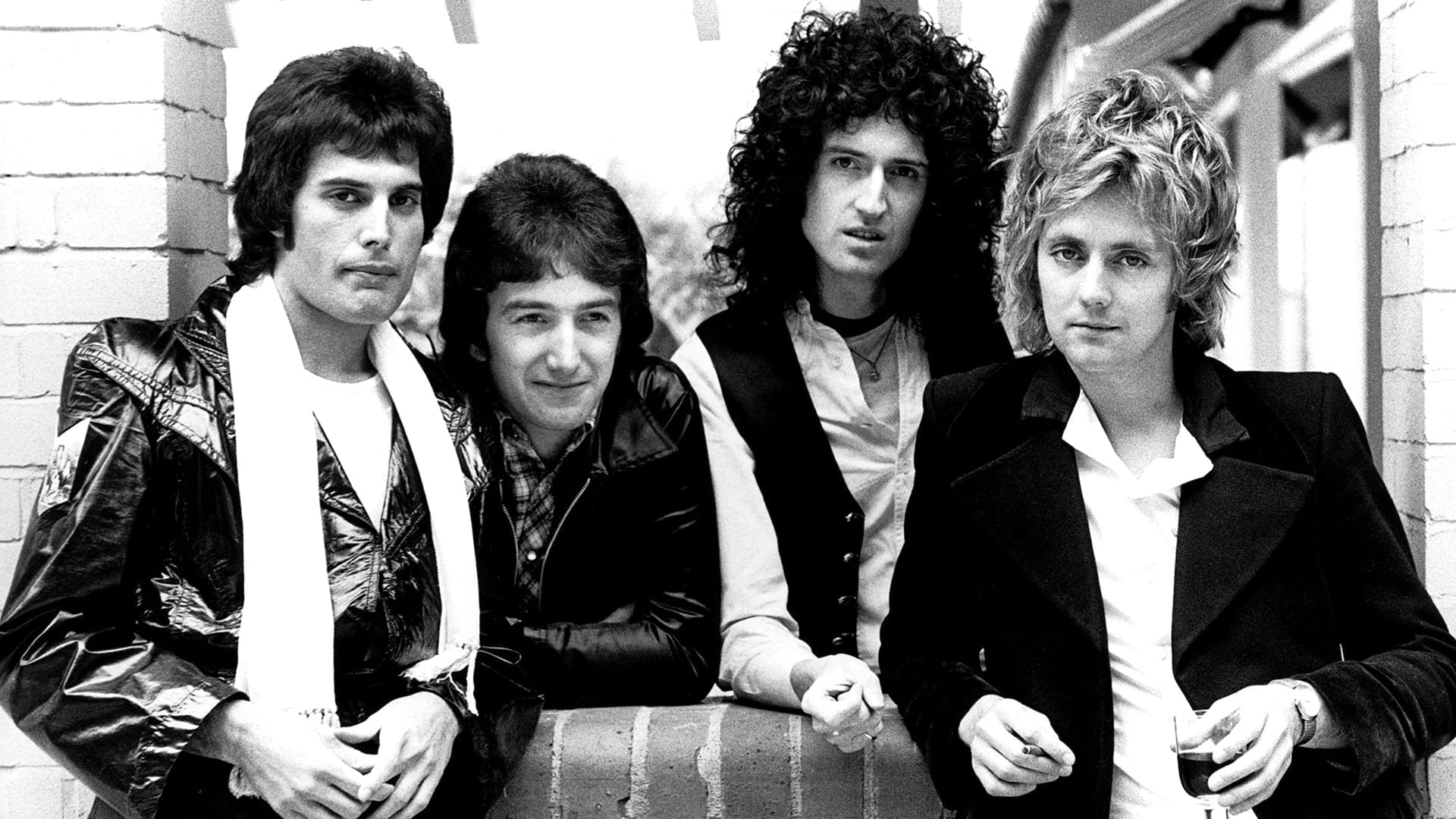 A black-and-white 1977 press photo of Queen: Freddie Mercury, John Deacon, Brian May, and Roger Taylor posed together leaning against a low wall.