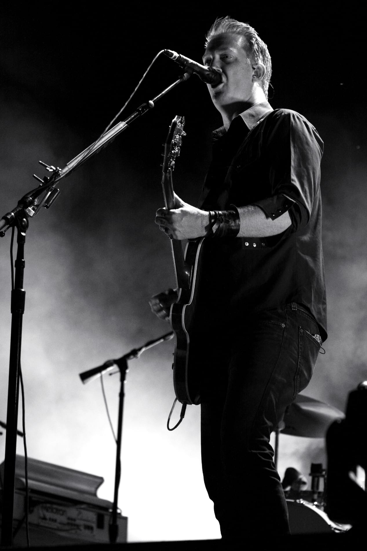 Josh Homme sings into a microphone while playing a guitar, backlit in a black and white stage photo.