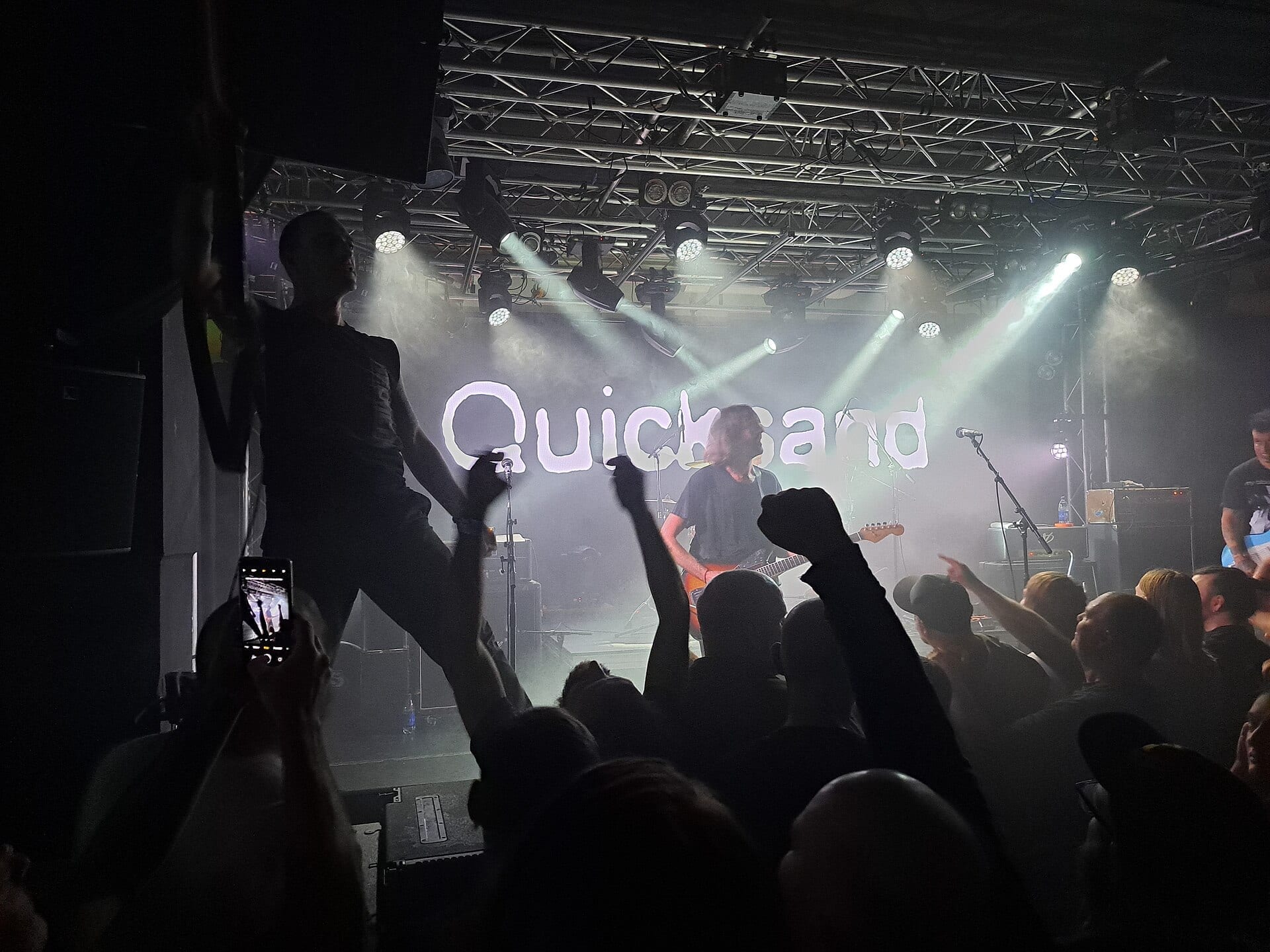 A silhouetted crowd raises fists in front of the stage as Quicksand performs beneath their illuminated band name and stage lights.