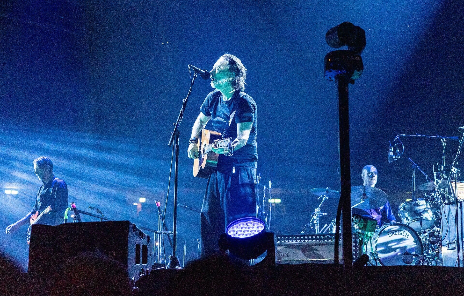 Thom Yorke sings into a microphone while playing acoustic guitar on stage under blue light, with bandmates on bass and drums behind him.