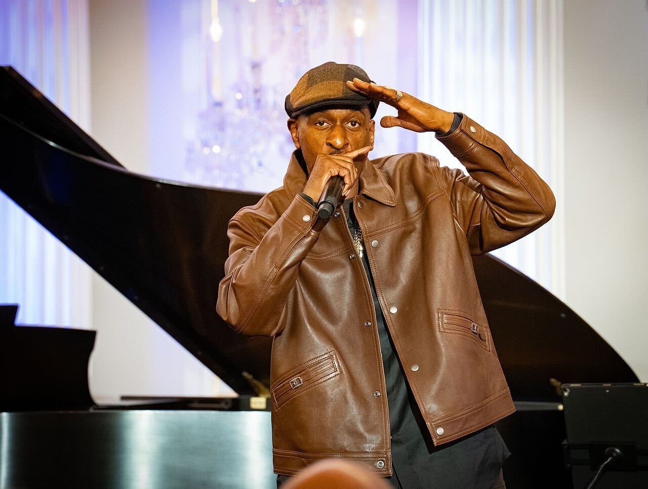 Rakim raises a hand to the brim of his flat cap while rapping into a microphone, standing beside a grand piano.