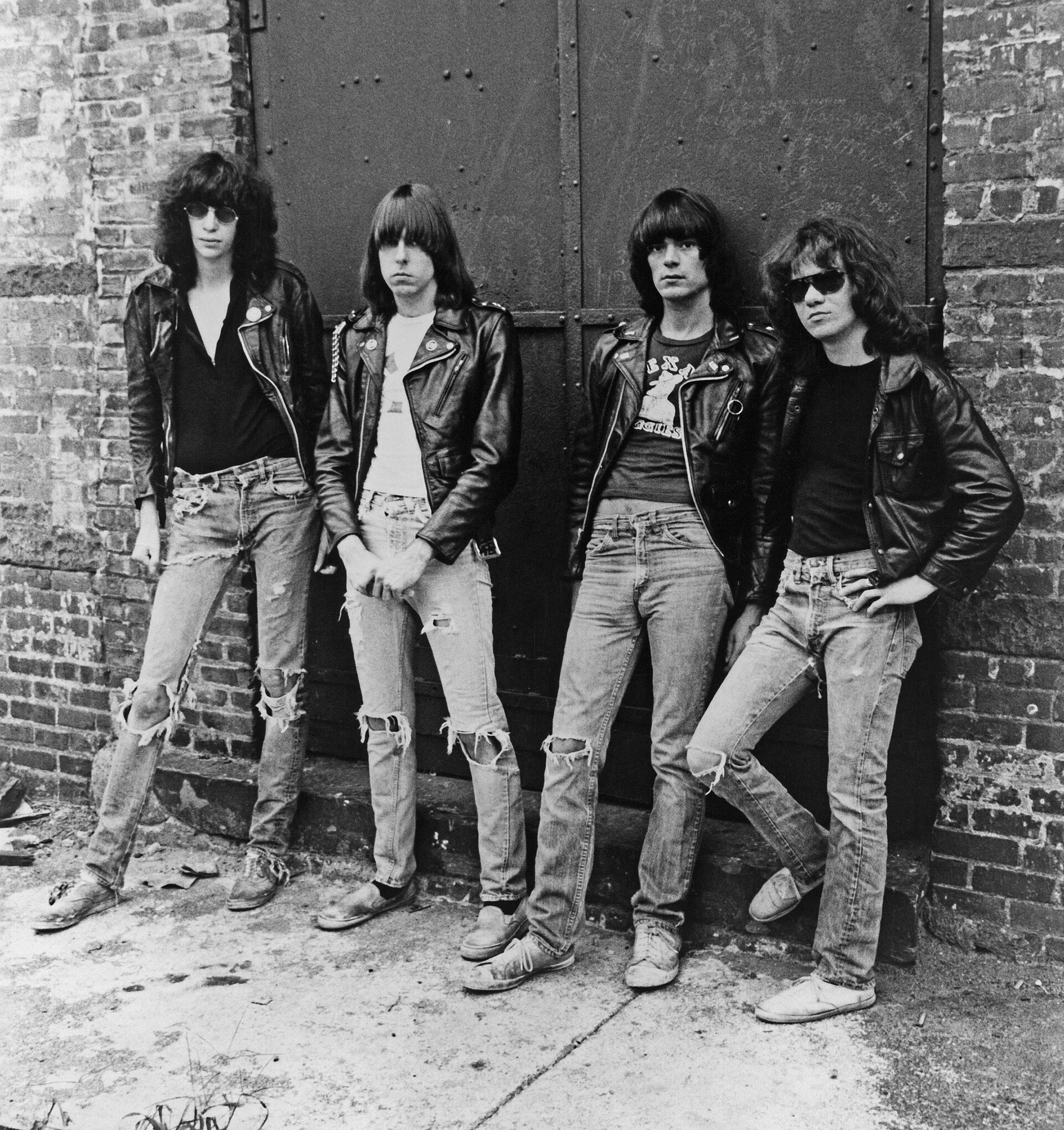 The four Ramones stand in a row against a brick wall in leather jackets, T-shirts and torn jeans, photographed in black and white.