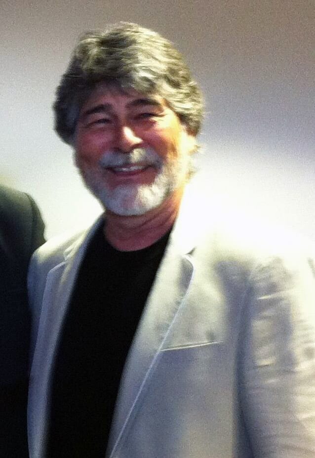Randy Owen, wearing a light gray blazer over a black shirt, smiles warmly in a close-up photo.