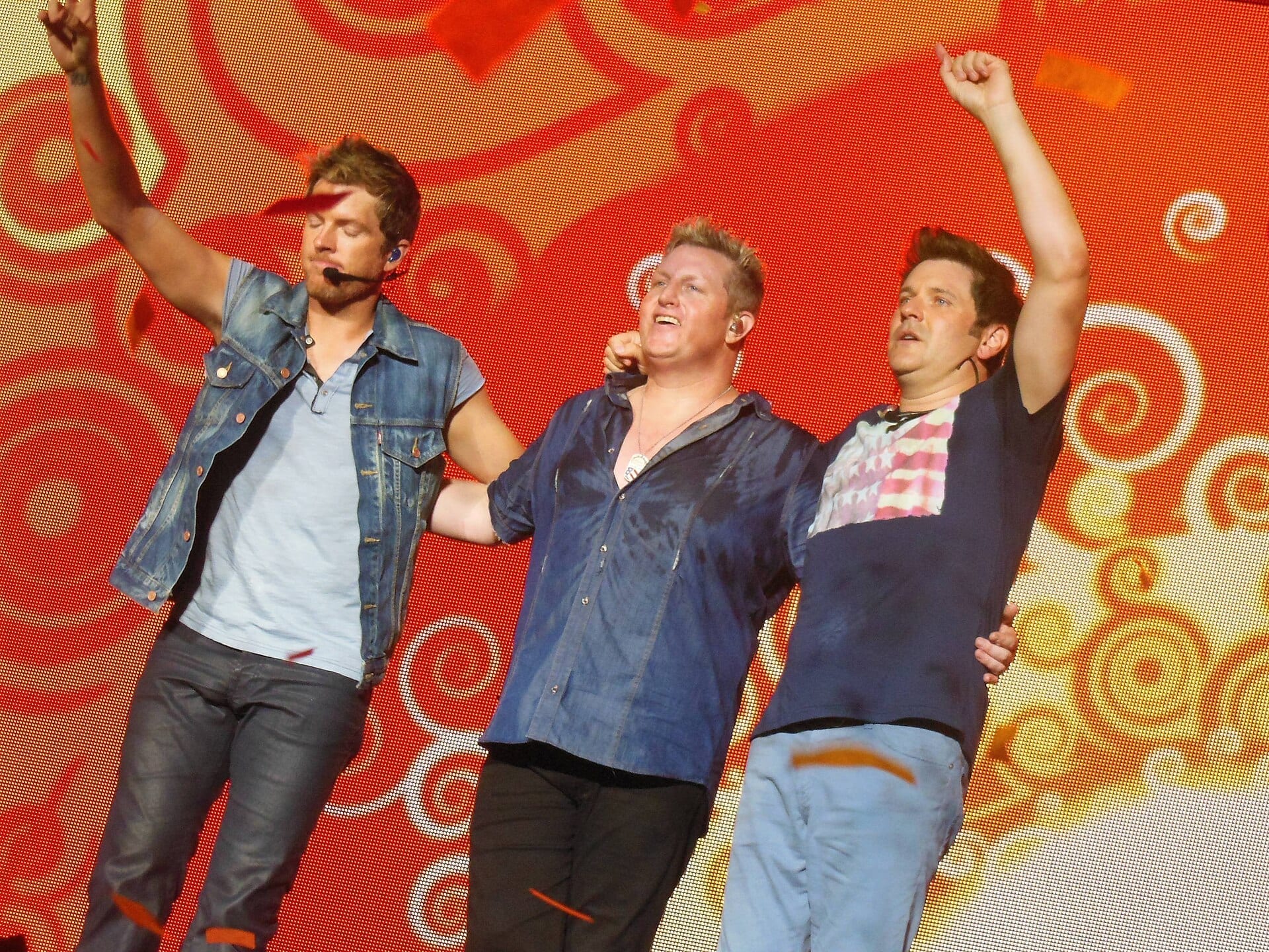 The three members of Rascal Flatts stand on stage with arms raised against a red and gold backdrop, one in a denim vest with a headset mic, one in an open blue shirt, and one in a red, white and blue tie-dye shirt.