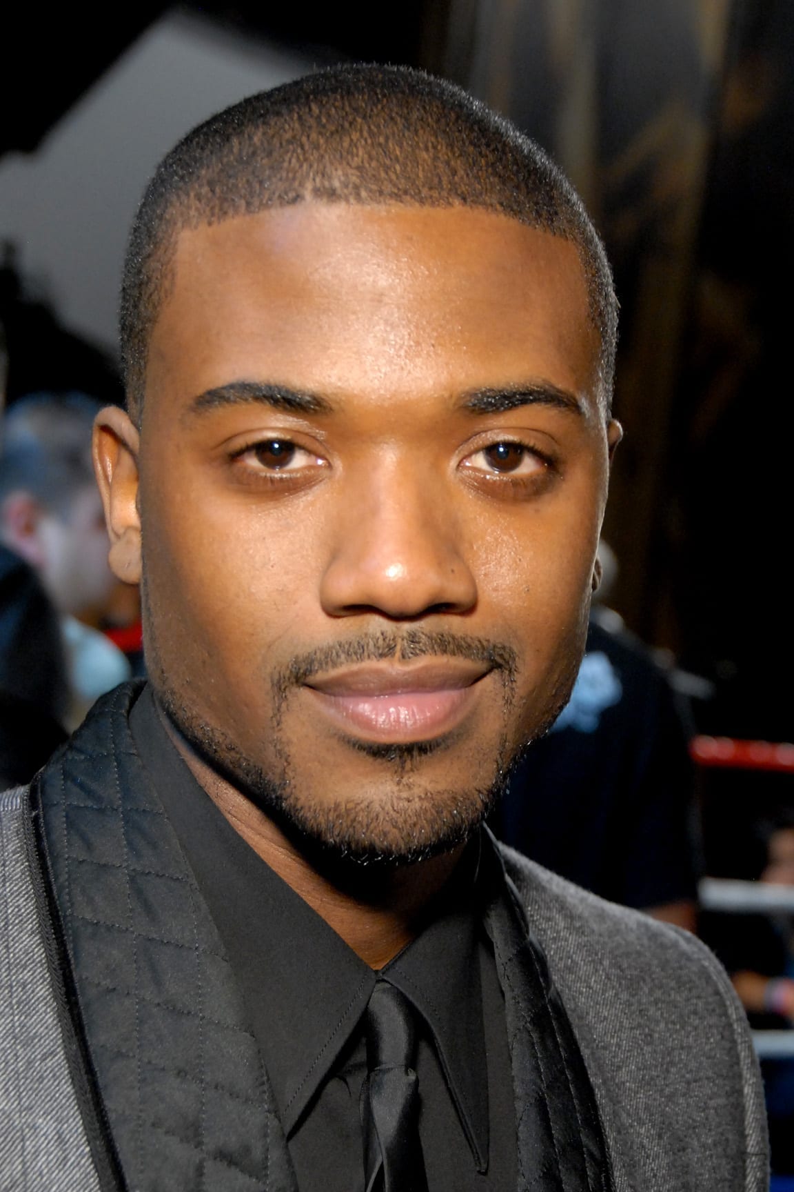 A close-up portrait of Ray J wearing a black shirt and tie under a gray blazer, with a neutral expression.