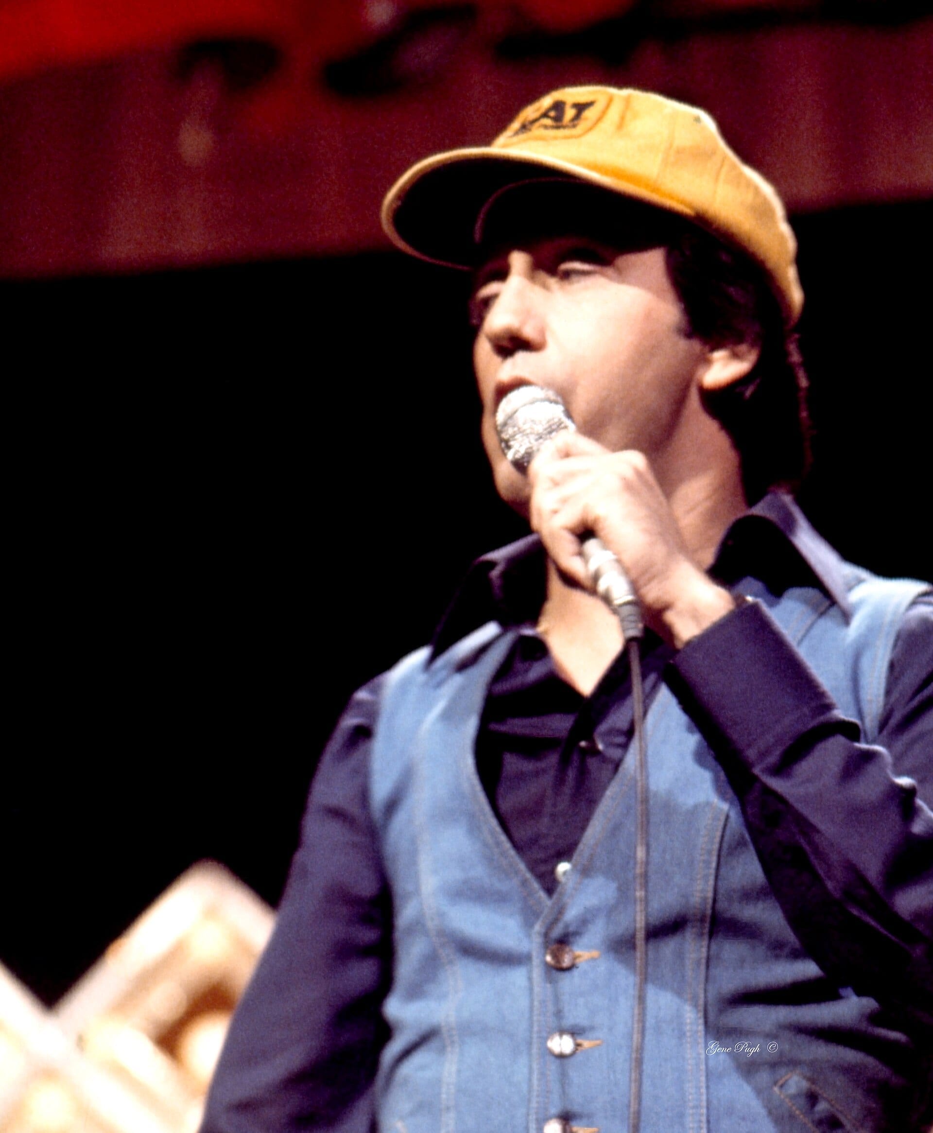Ray Stevens, wearing a yellow cap and a denim vest over a dark shirt, sings into a handheld microphone.